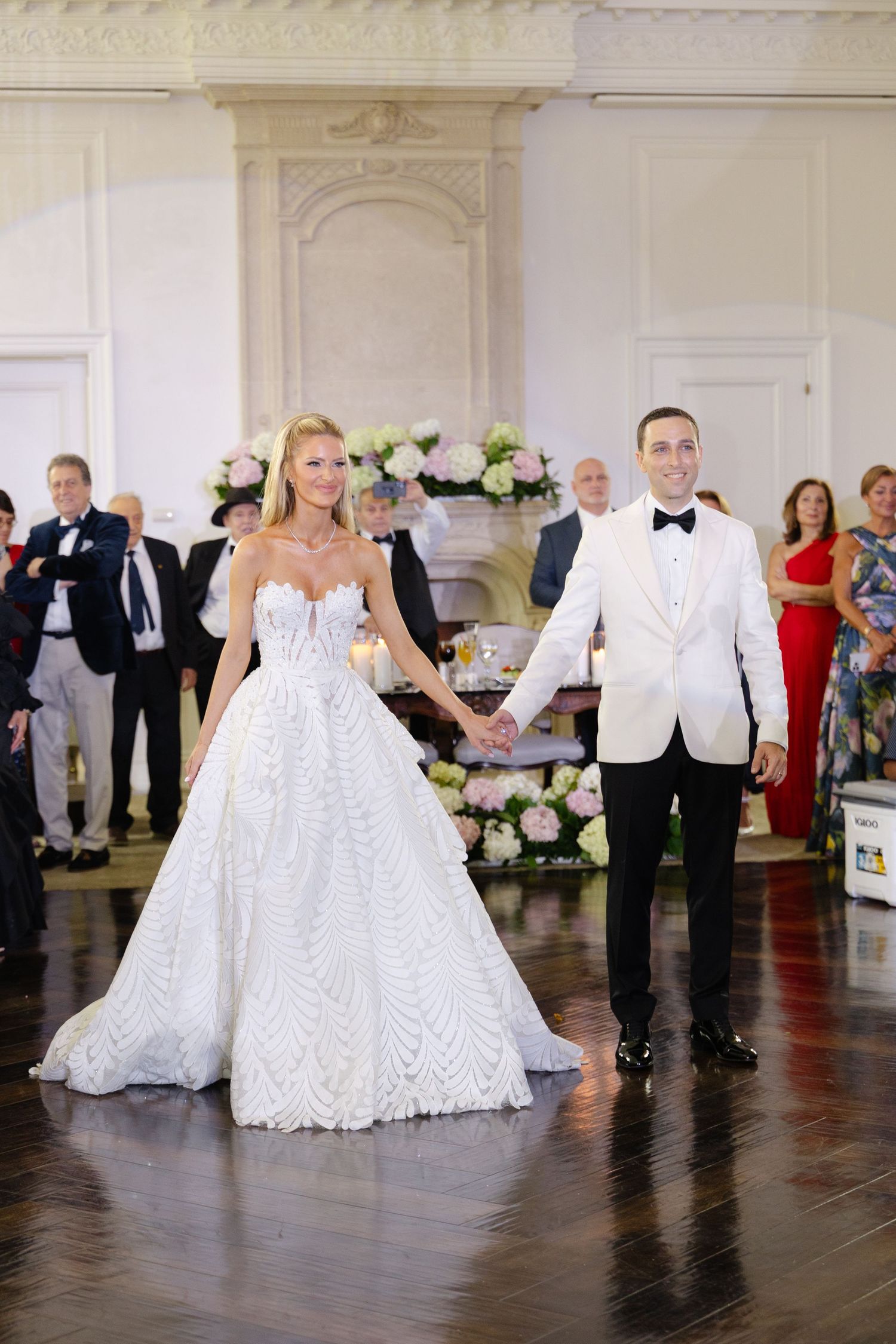 Newlyweds hold hands during their first dance in an elegant white ballroom decorated with floral arrangements.