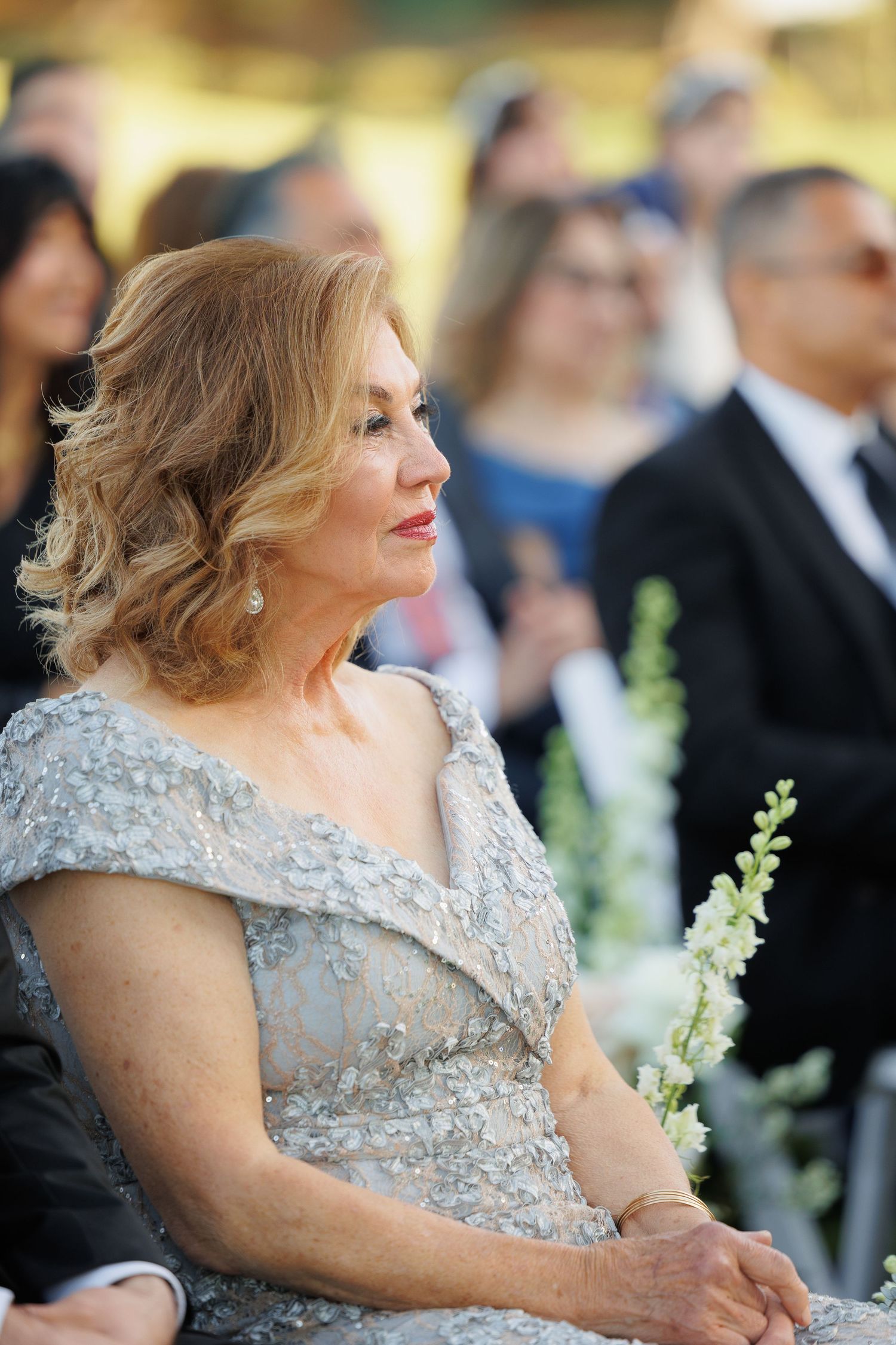 Woman in elegant silver beaded evening gown sits among guests at formal outdoor event.