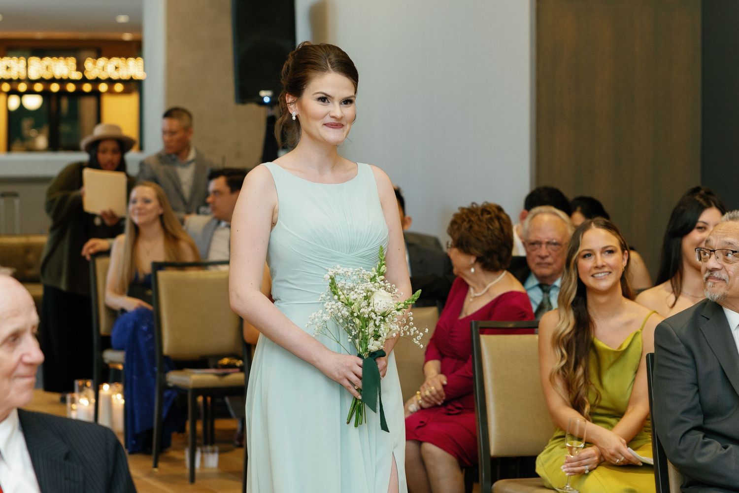 Wedding party members in formal attire stand together holding bouquets at an indoor ceremony.