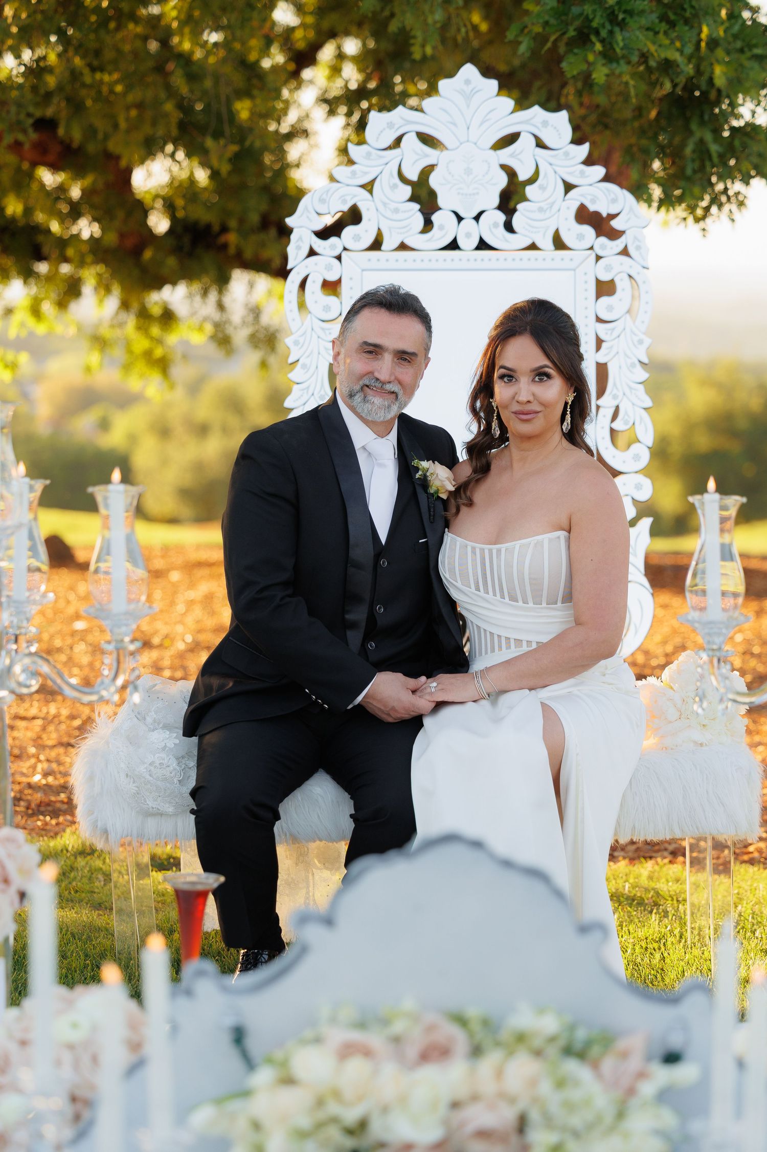 A romantic wedding couple poses on white vintage chairs surrounded by autumn leaves and sunlit trees in an outdoor setting.
