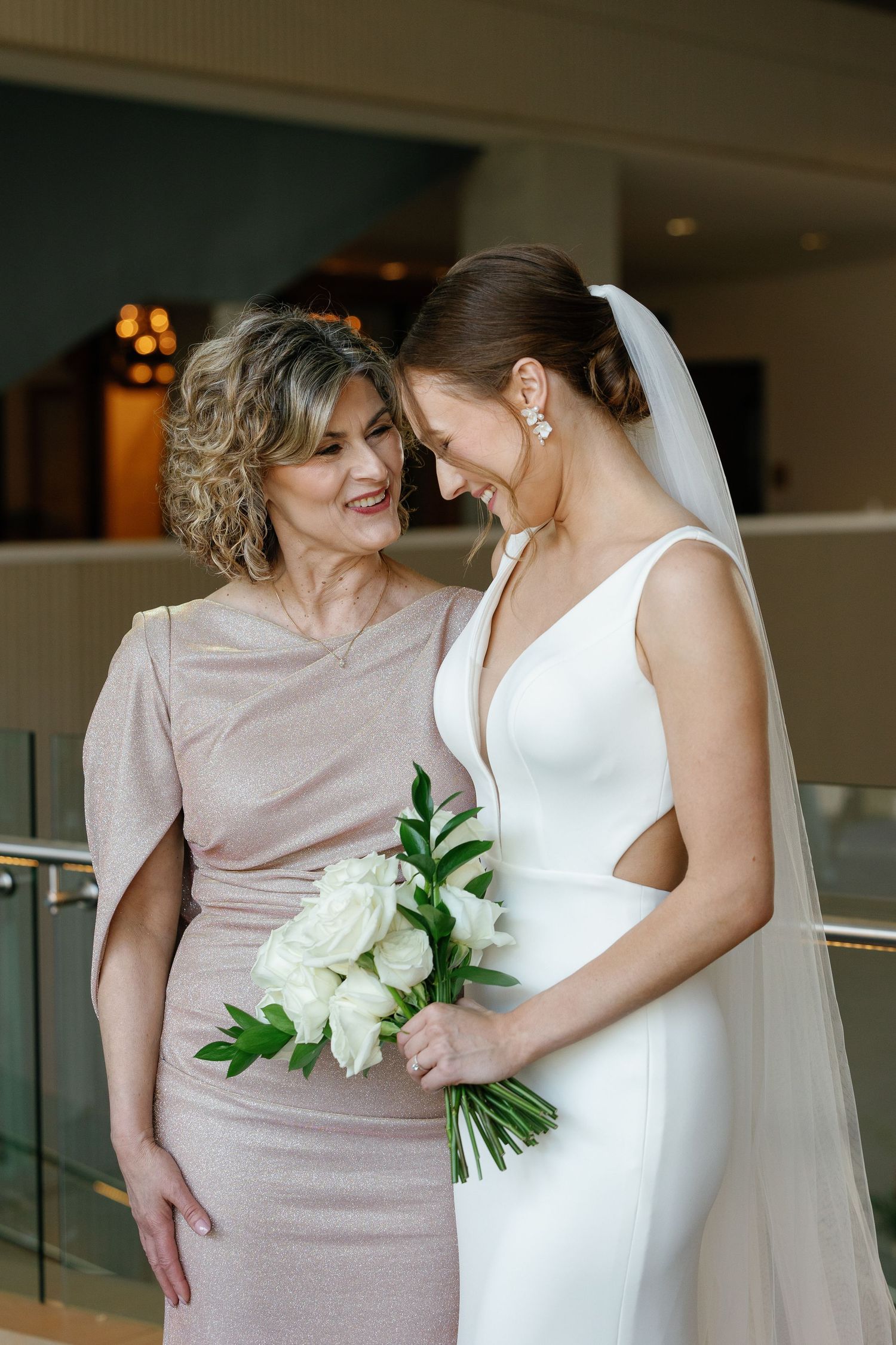 Mother and bride share an intimate moment in elegant blush and white gowns with white floral bouquet.