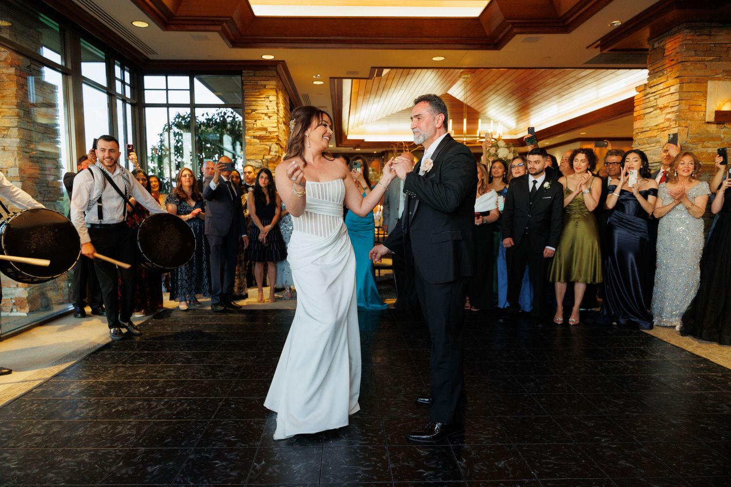 A couple shares their first dance on a dark dance floor while wedding guests cheer and applaud in the background.