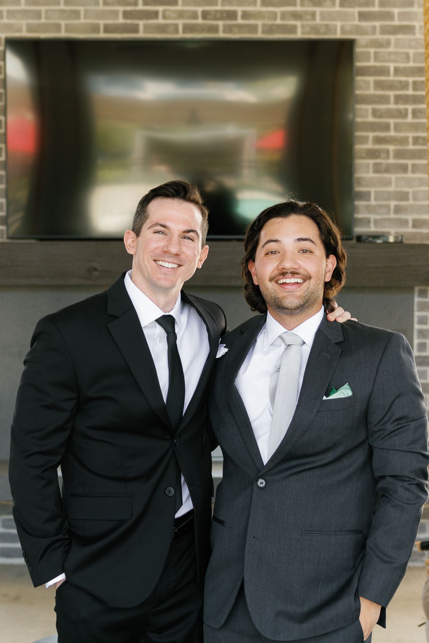 Two men in formal black suits and ties share a candid moment while posing together at a brick wall backdrop.