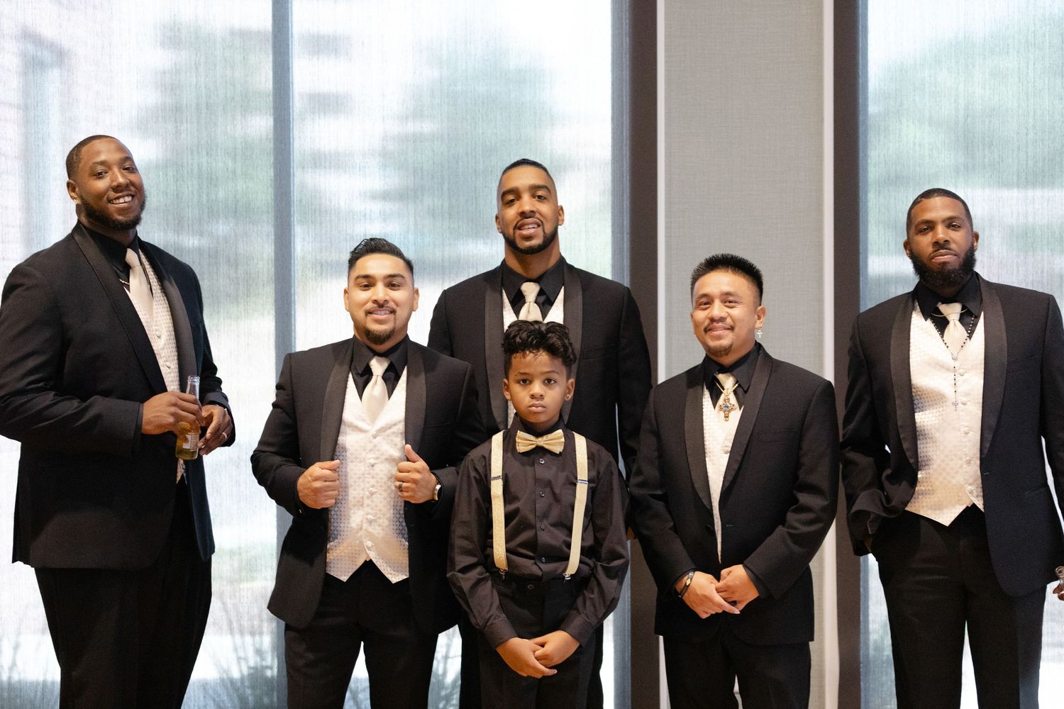 A formal wedding party wearing black tuxedos and suits stands in a row for a group photo.