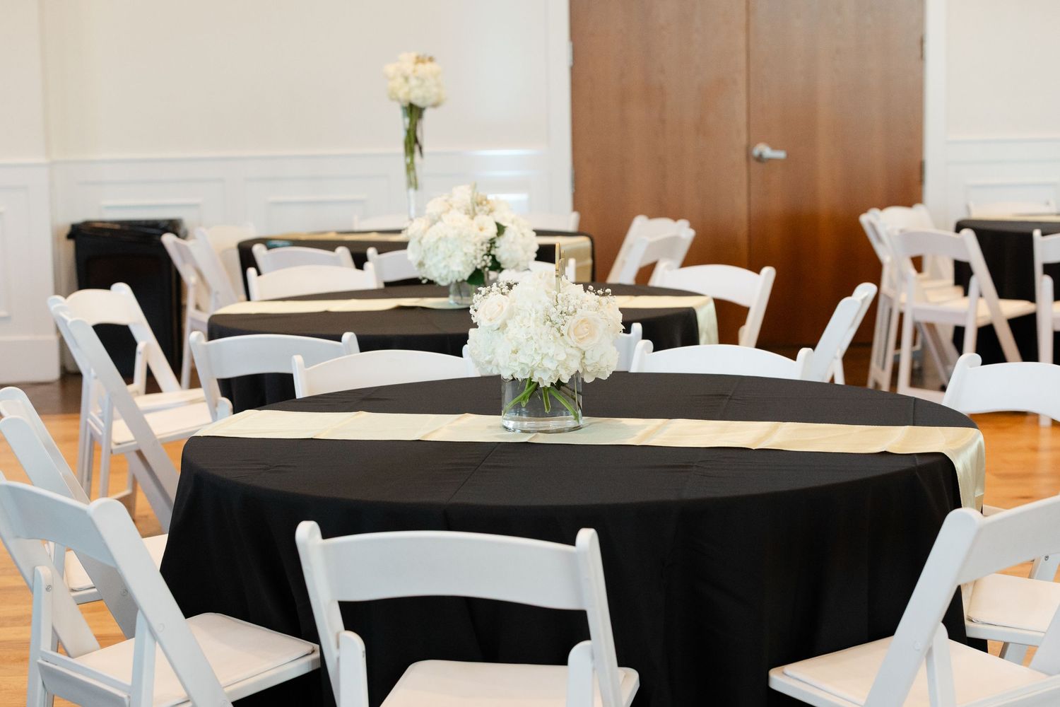 A formal event space with round tables covered in black tablecloths and white floral centerpieces.