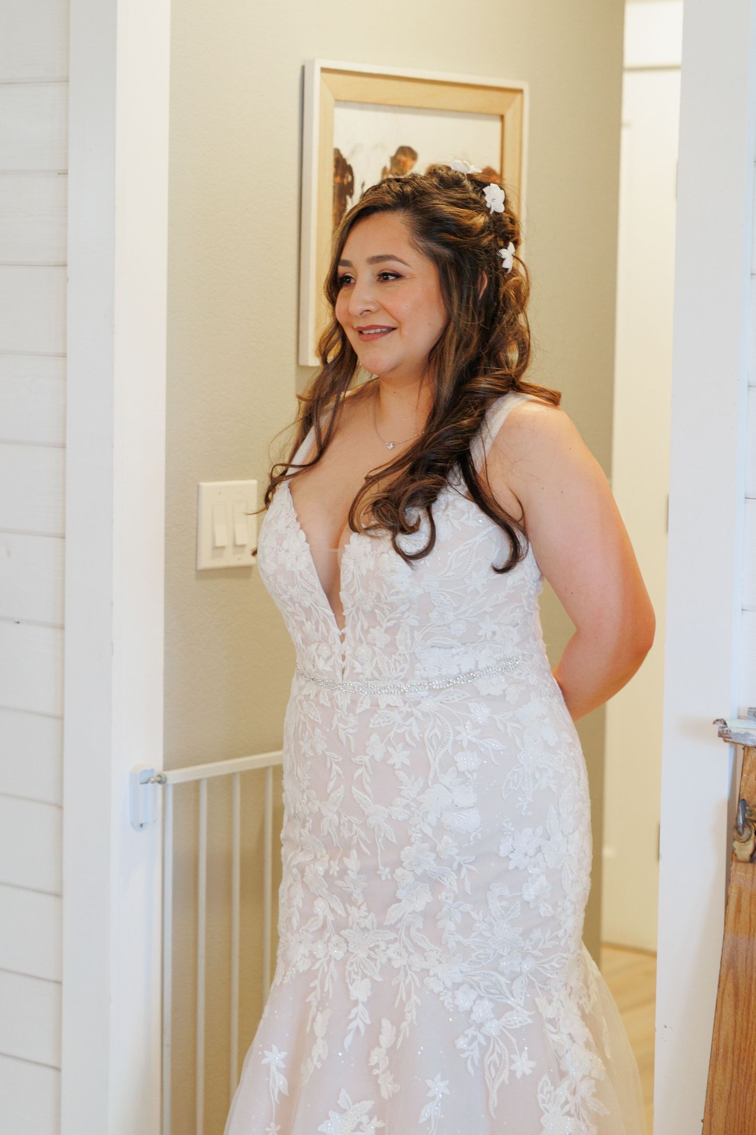 A bride in a white lace V-neck wedding gown smiles while getting ready in a bright interior space.