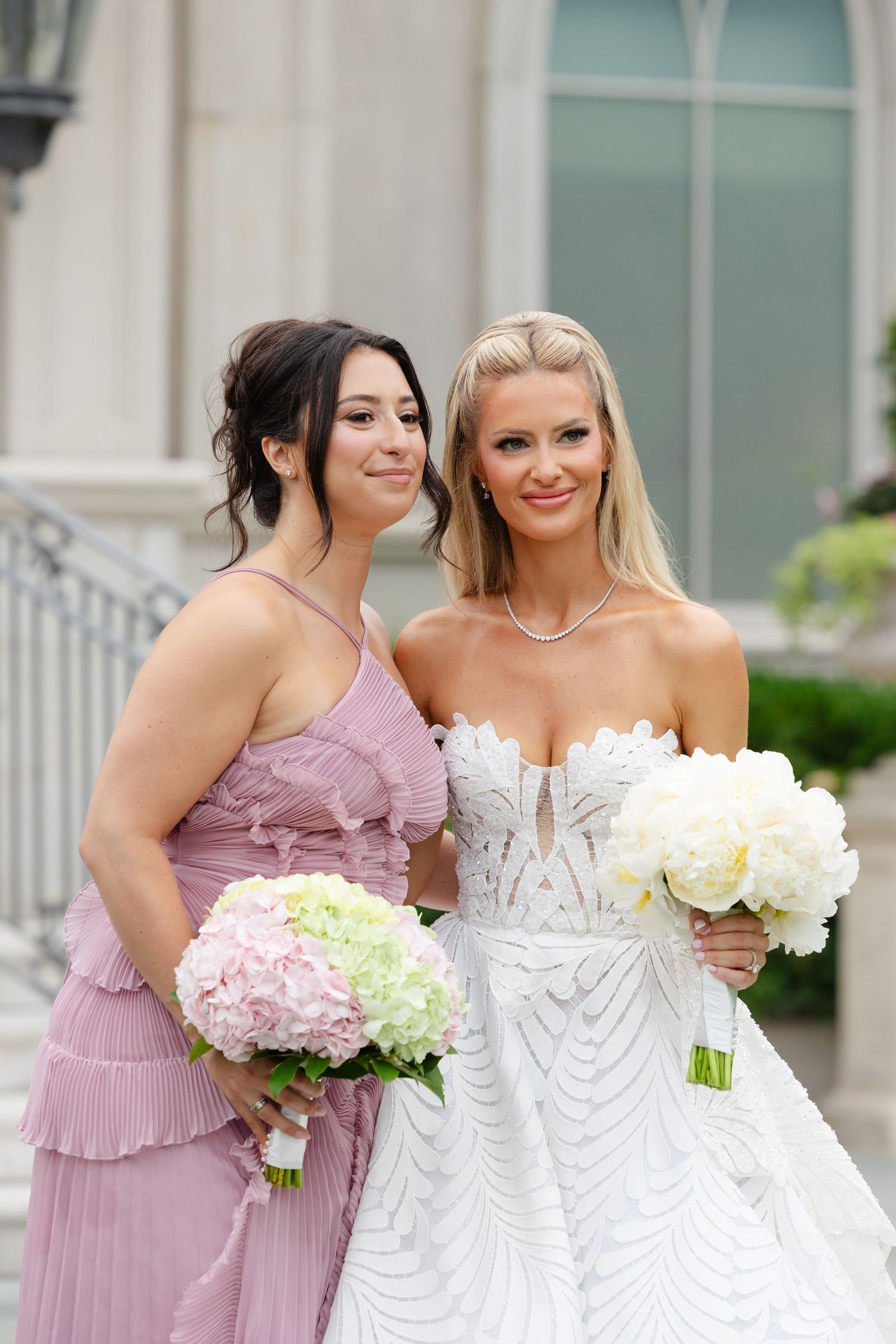 A bride in a white beaded gown and bridesmaid in dusty rose dress hold pink and white hydrangea bouquets.