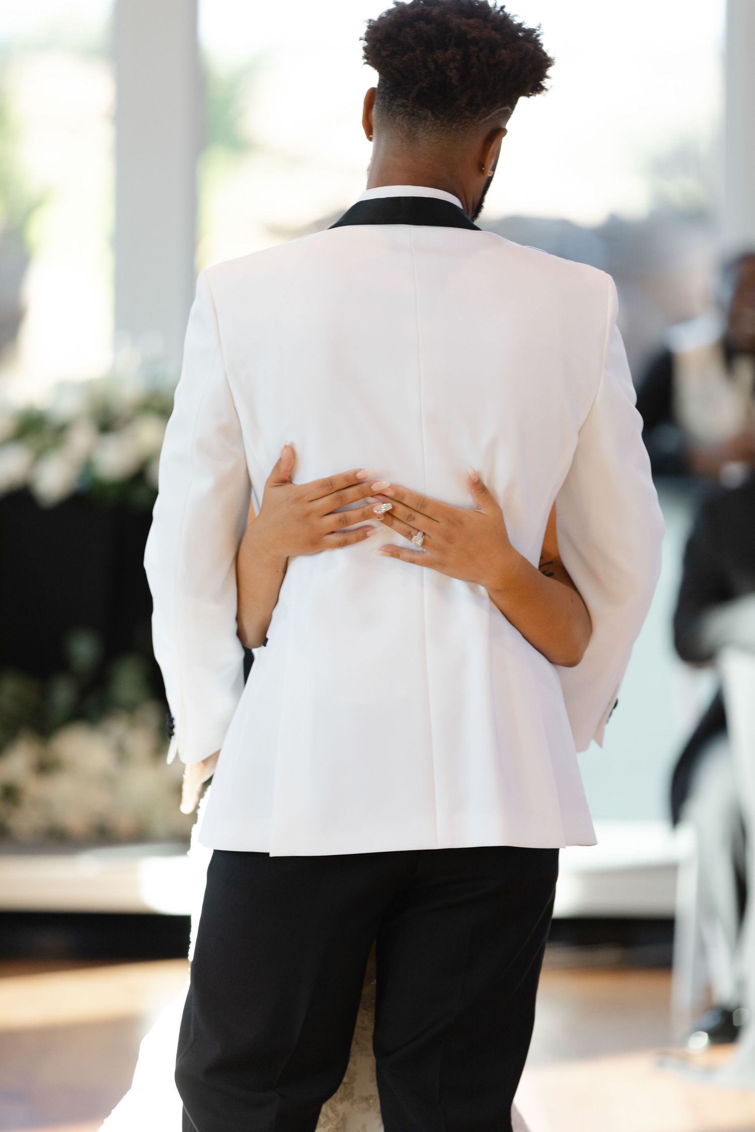 A couple embraces during a wedding dance, with hands wrapped around a white tuxedo jacket.