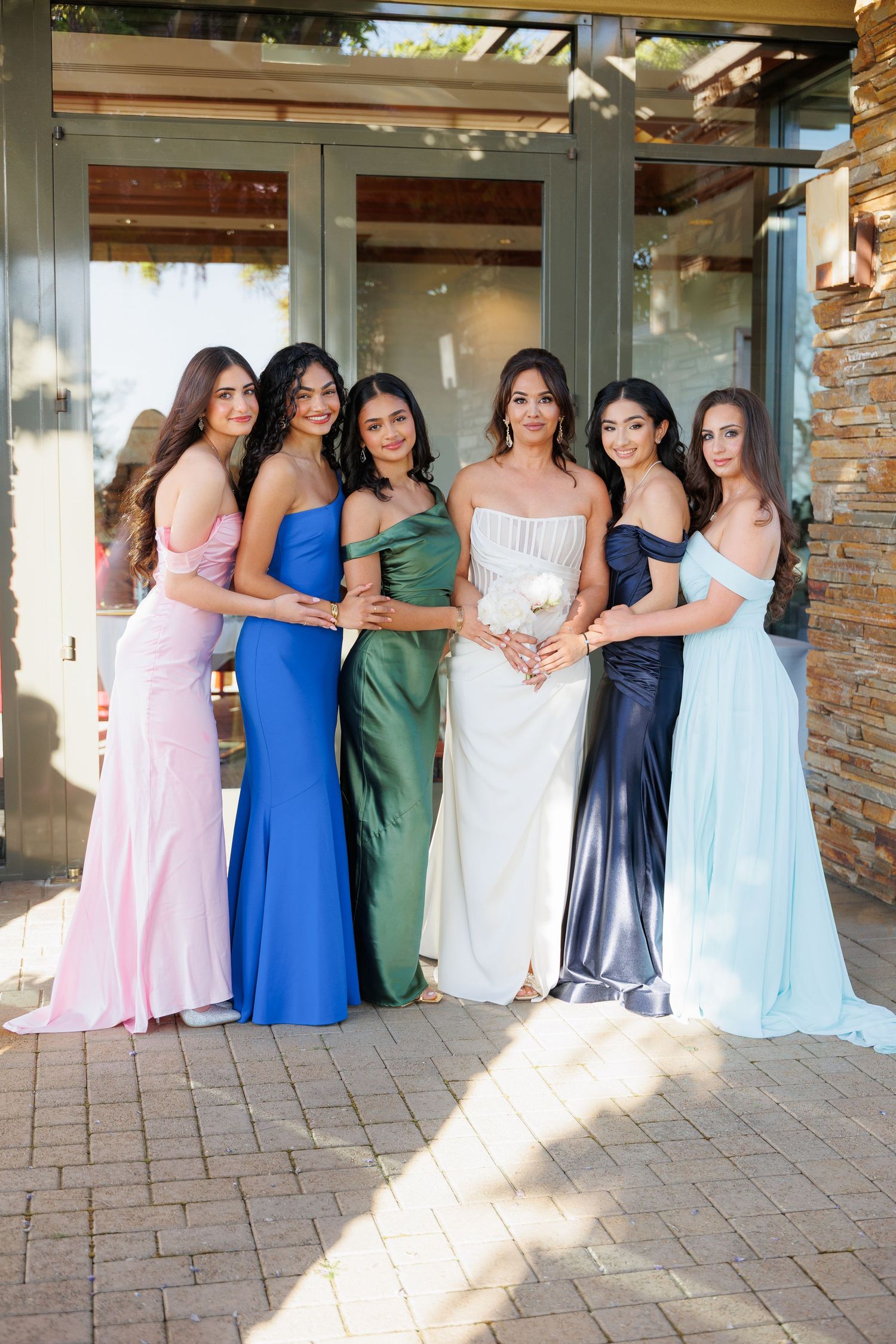 A group in formal dresses poses together on a patio with brick walls and sunlight streaming in.