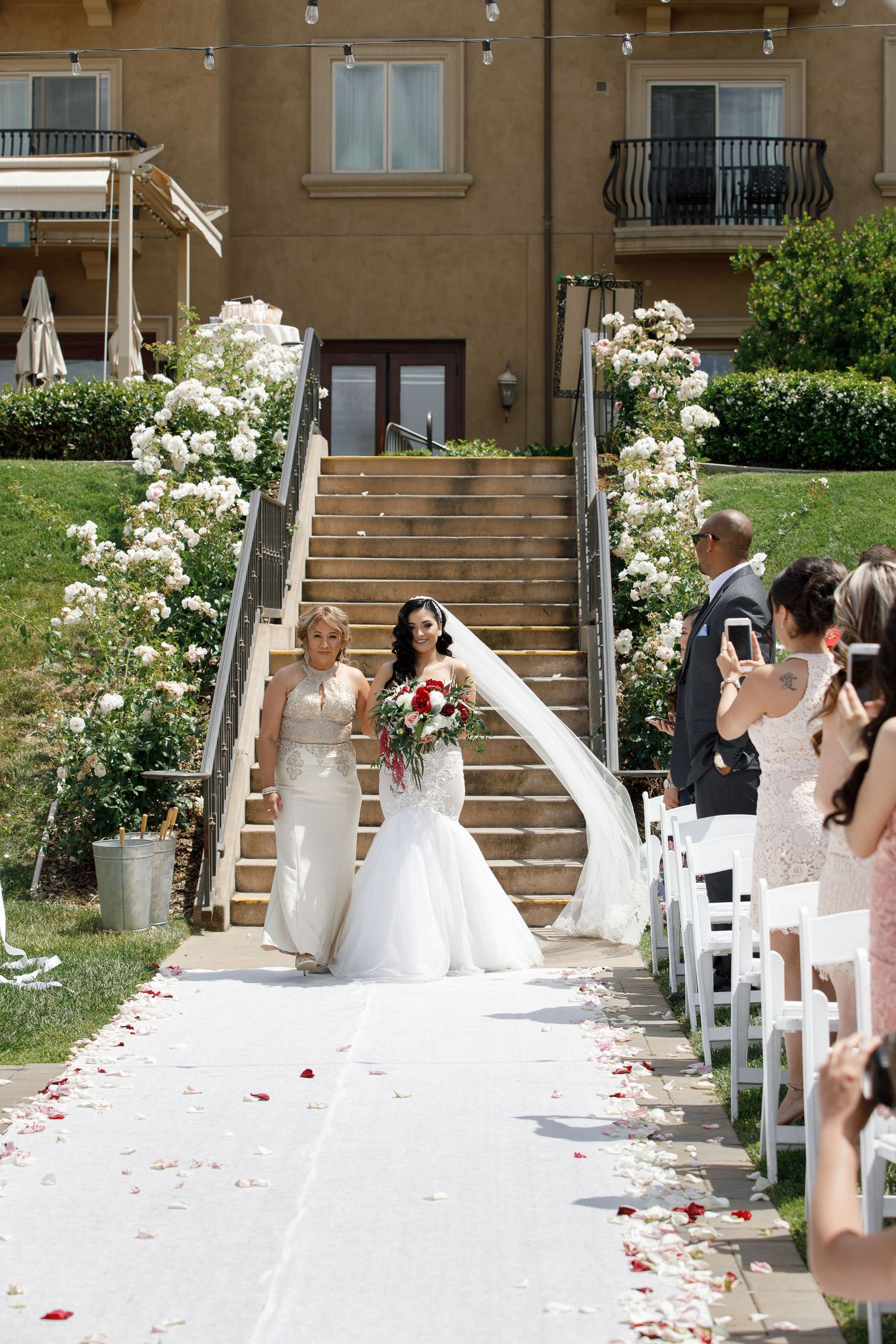 A wedding ceremony setup with white chairs and rose petals scattered along an outdoor staircase leading to a building entrance.