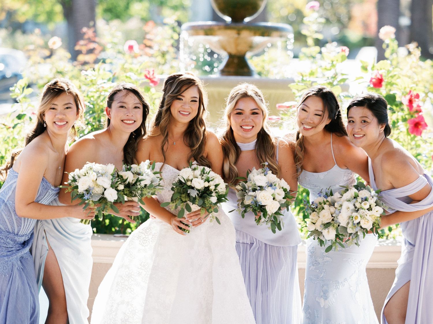 A bridal party in light blue and white dresses holds white floral bouquets while laughing together in a garden setting.