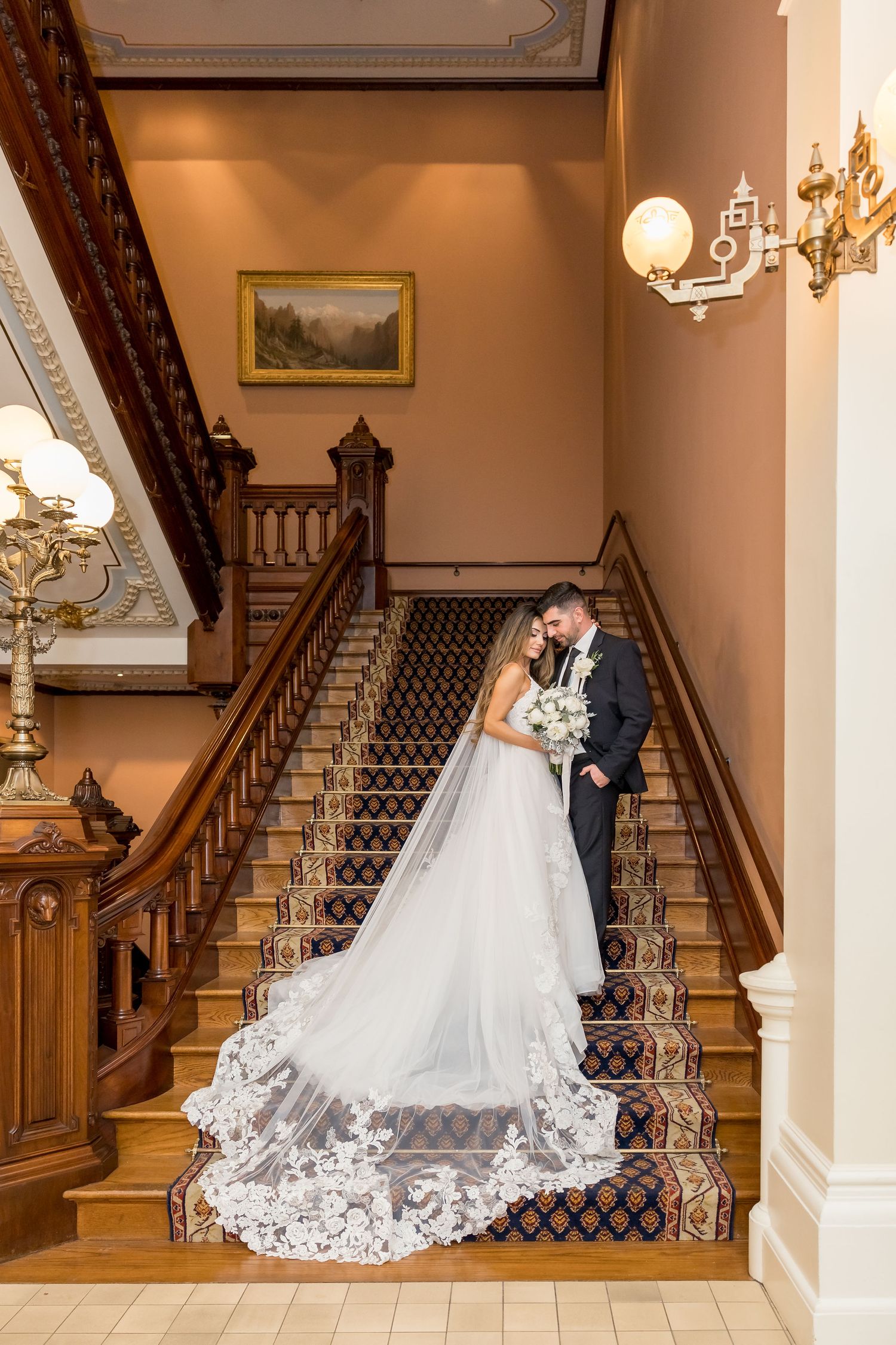 Elegant wedding couple photographed on grand wooden staircase with ornate carpet runner in historic mansion interior.