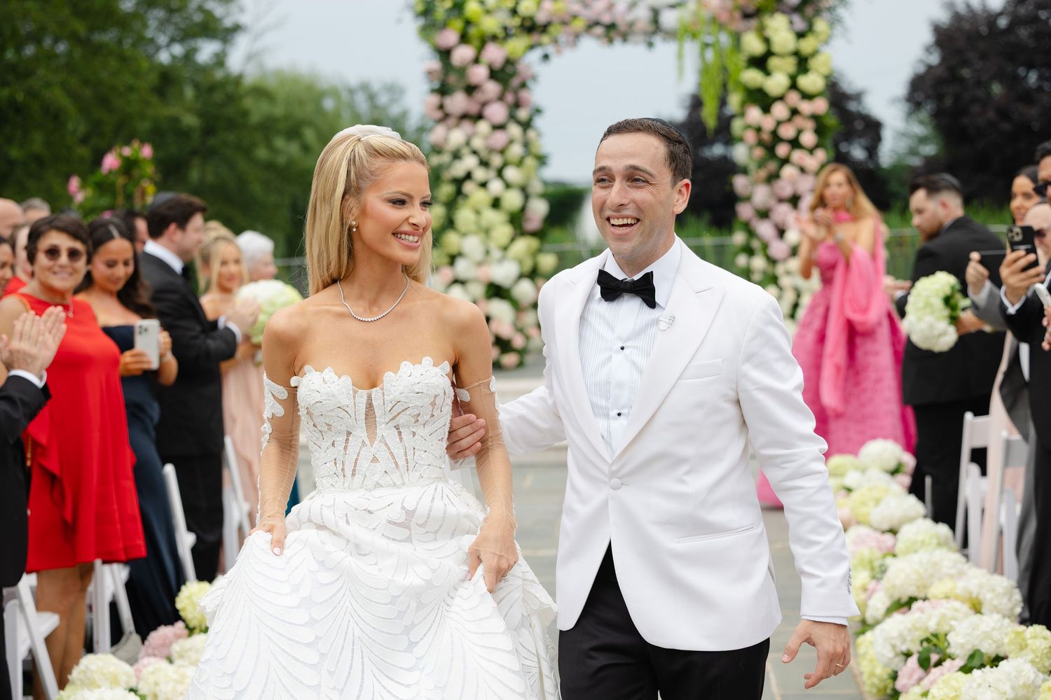Bride in strapless beaded gown and groom in white tuxedo jacket walk joyfully down the aisle at outdoor wedding.