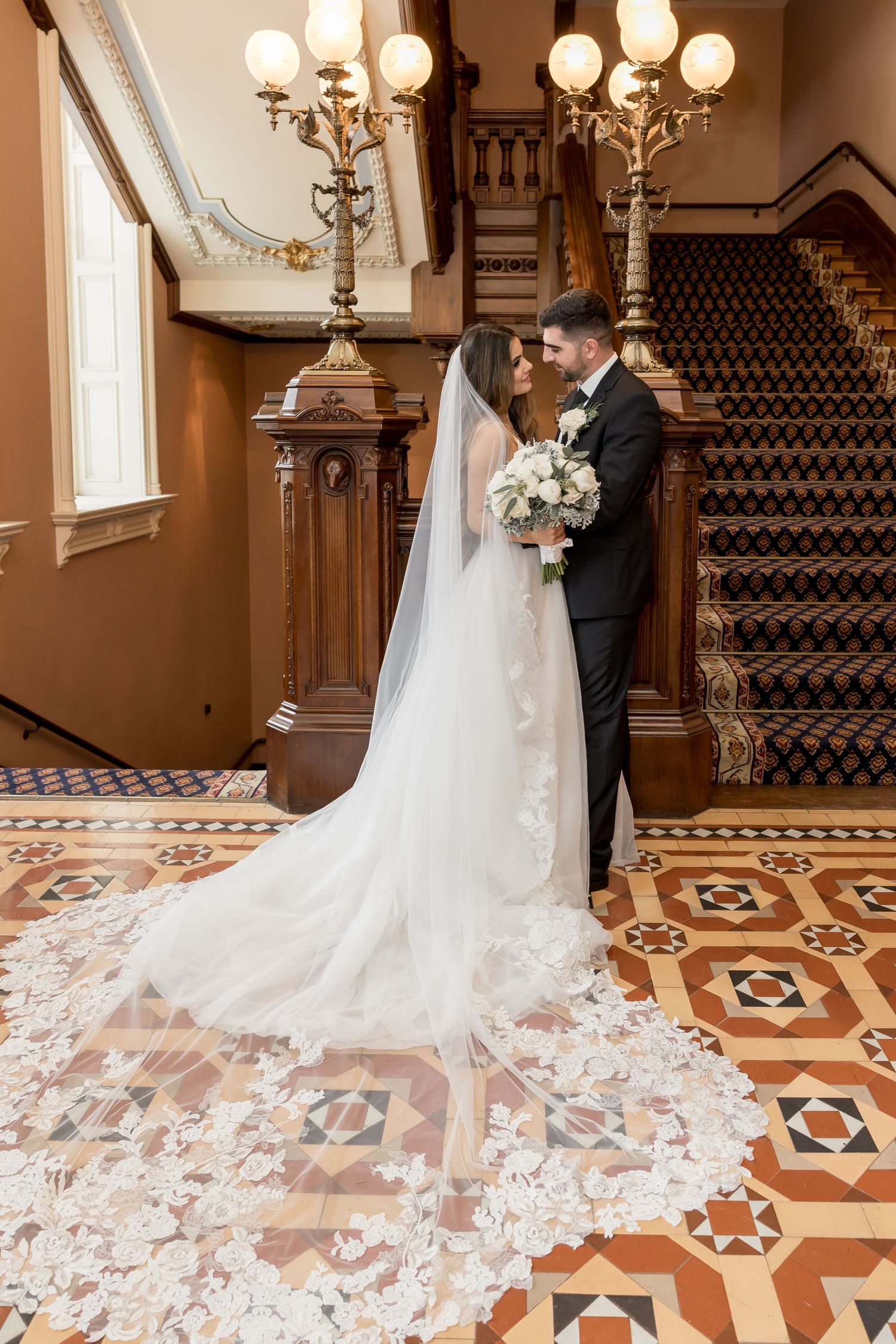Wedding couple stands in an elegant foyer with ornate tiled floors and a grand staircase with flowing lace veil details.