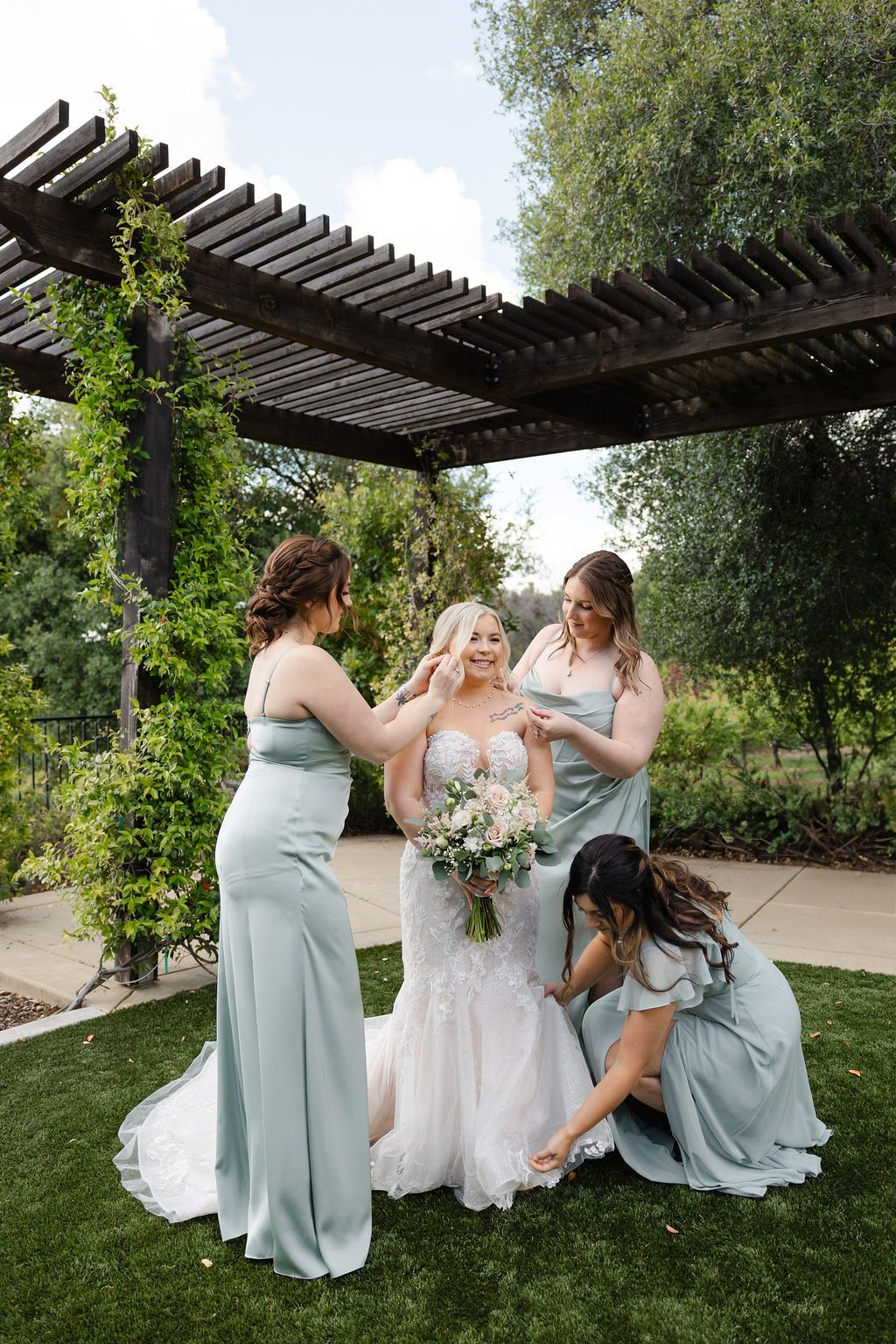 Wedding party in sage green dresses gathered under wooden pergola in garden setting.