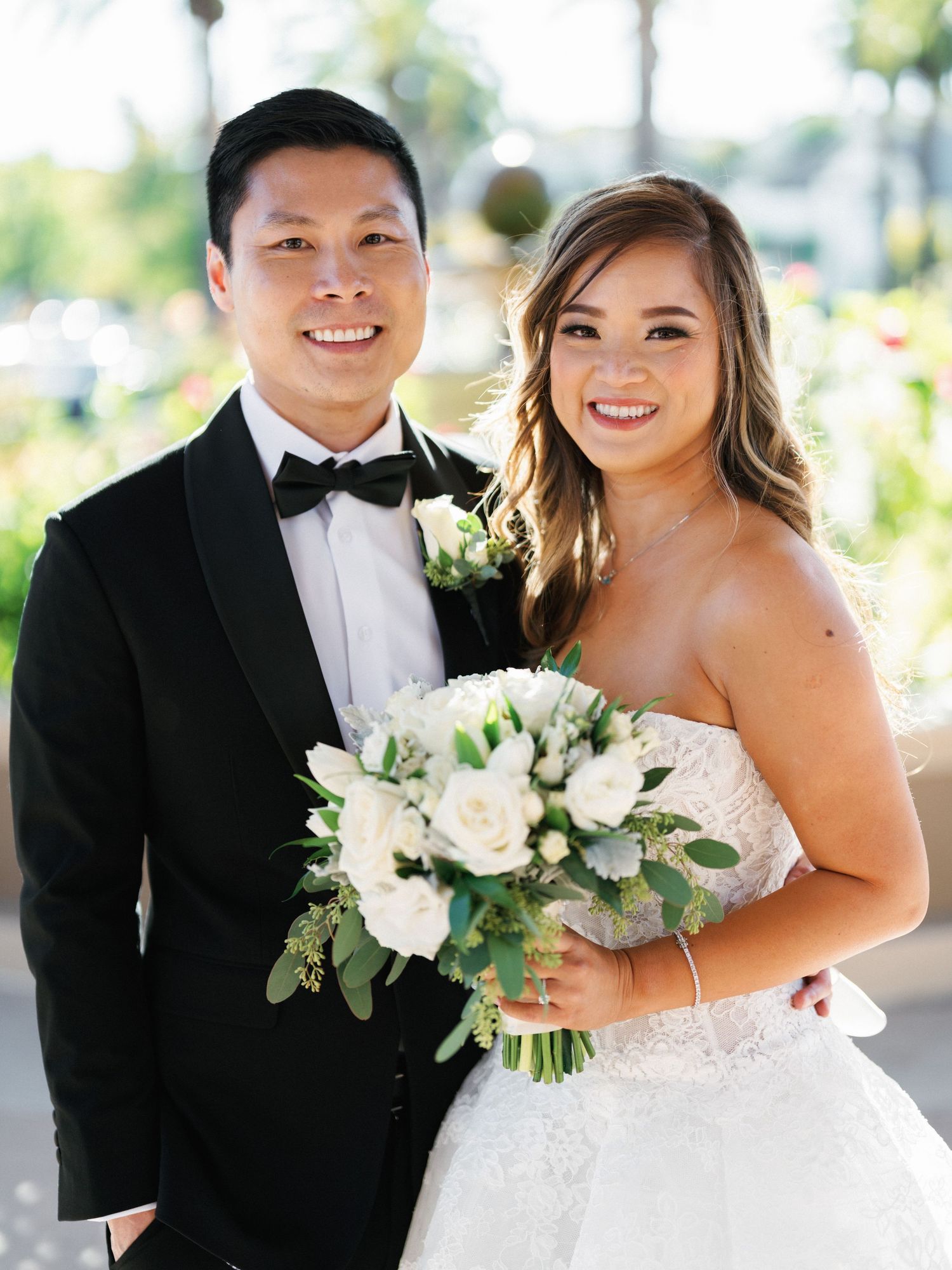 A bride and groom pose together outdoors, with the bride holding a white rose and greenery bouquet.