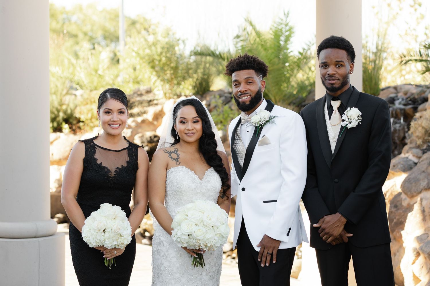 A wedding party in formal black and white attire poses together in an elegant outdoor setting with white columns.
