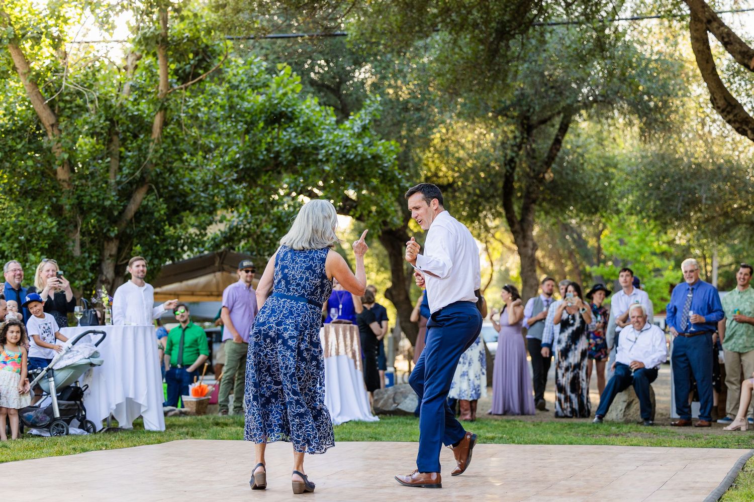 Wedding guests dancing in a line at an outdoor garden reception surrounded by lush greenery and trees.