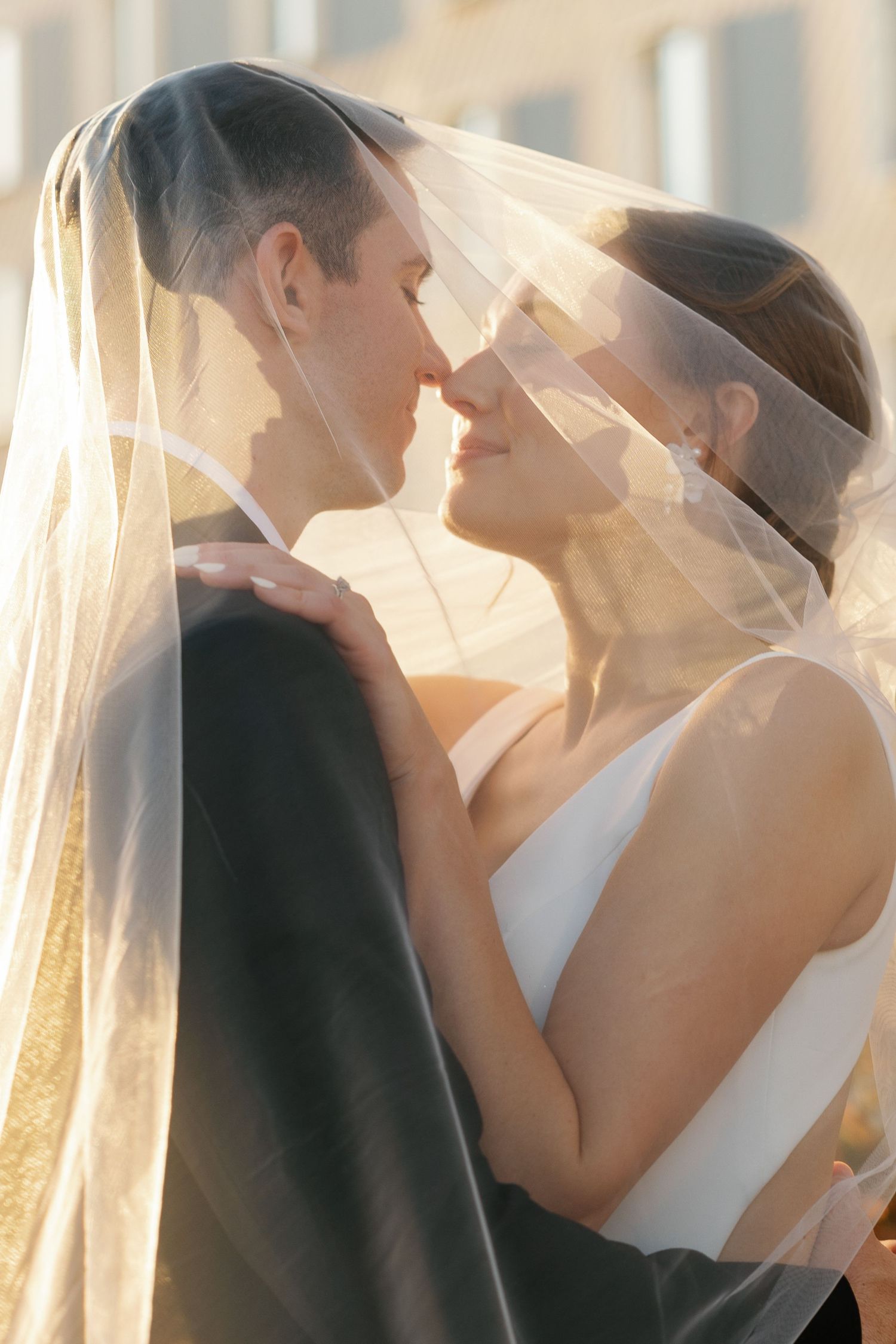 Romantic wedding couple embraces under flowing veil during golden hour sunset.