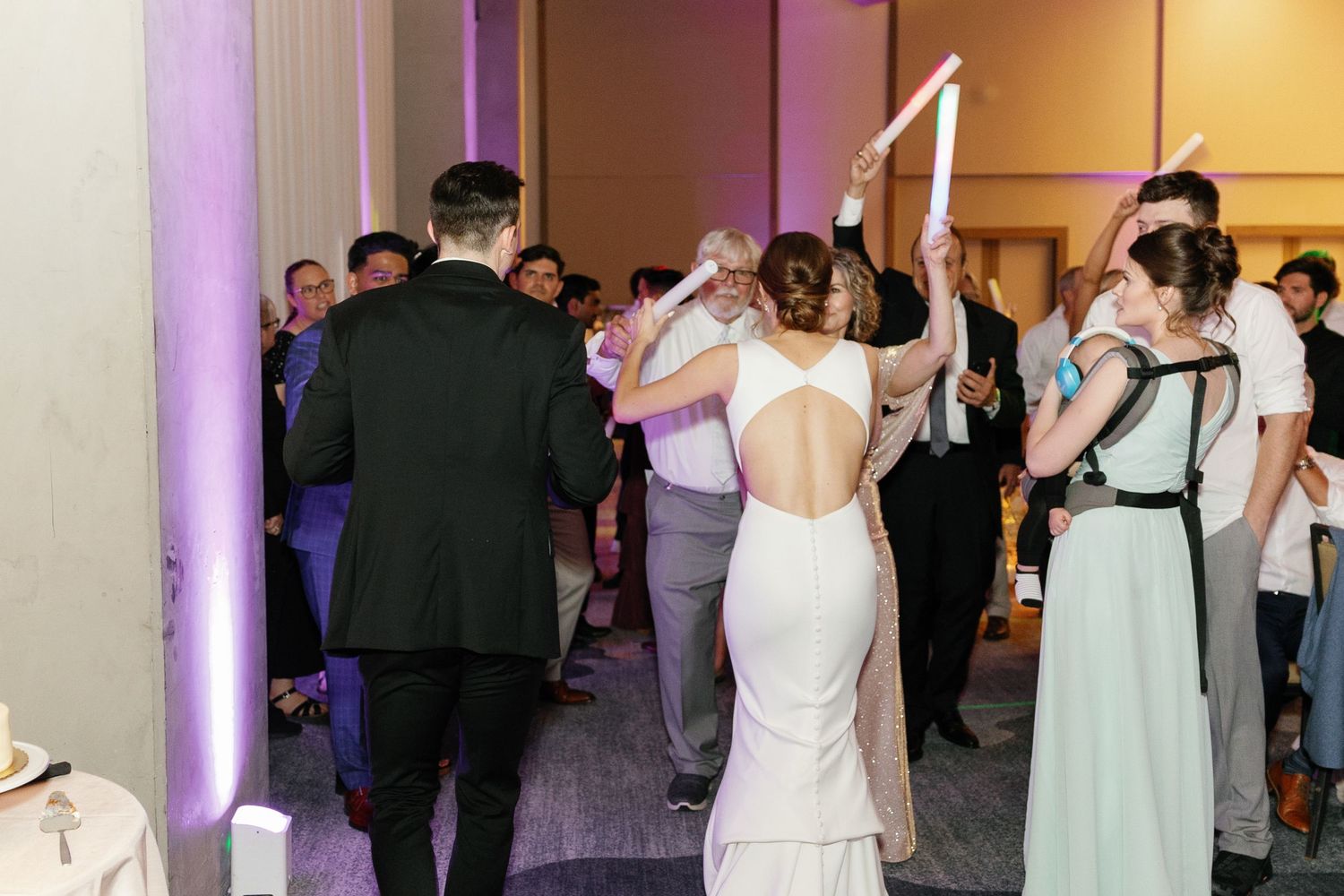 Wedding guests dance together on a dimly lit dance floor with pink and purple lighting at an elegant reception venue.