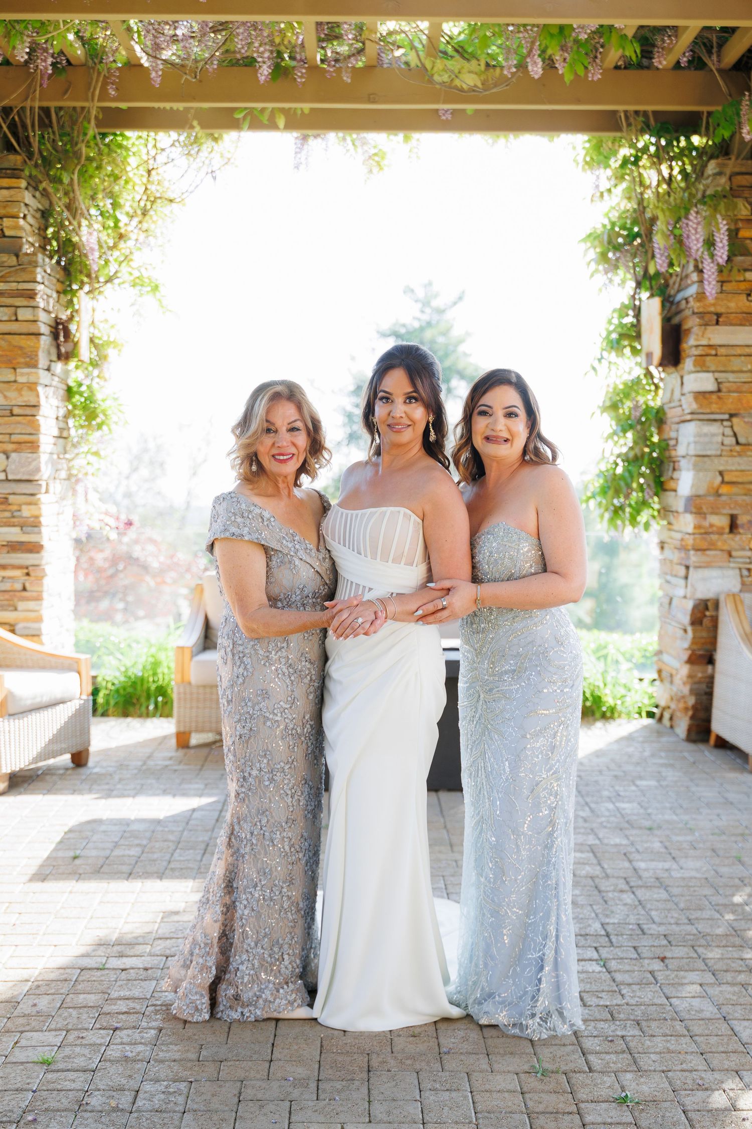 Bridesmaids in sparkly silver gowns pose with bride in white dress on outdoor patio with stone columns and greenery.