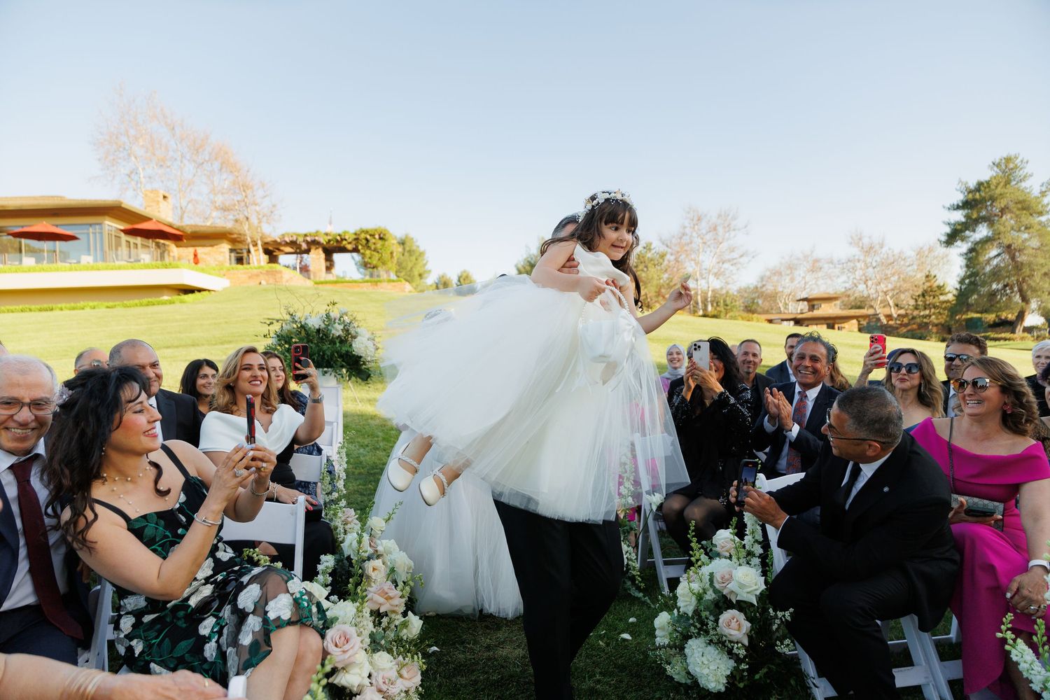 Wedding guests gather outdoors for a celebration with parasols and umbrellas under a bright blue sky.