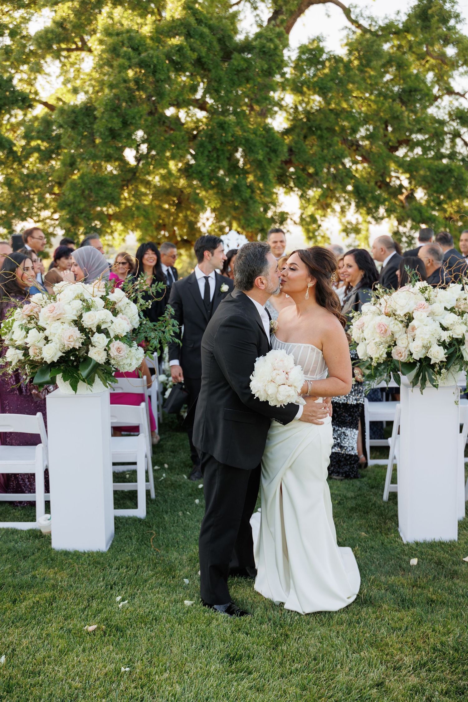 A romantic kiss between newlyweds at an outdoor garden wedding ceremony with white floral arrangements and folding chairs.