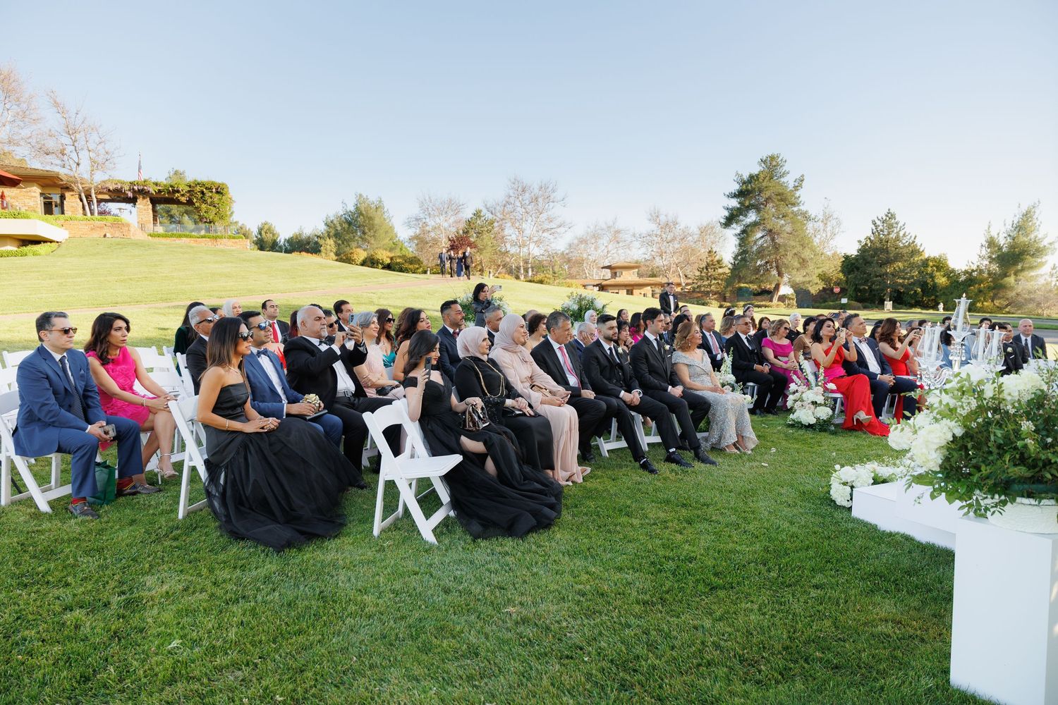 Wedding guests seated in white chairs on a green lawn during an outdoor ceremony at sunset.