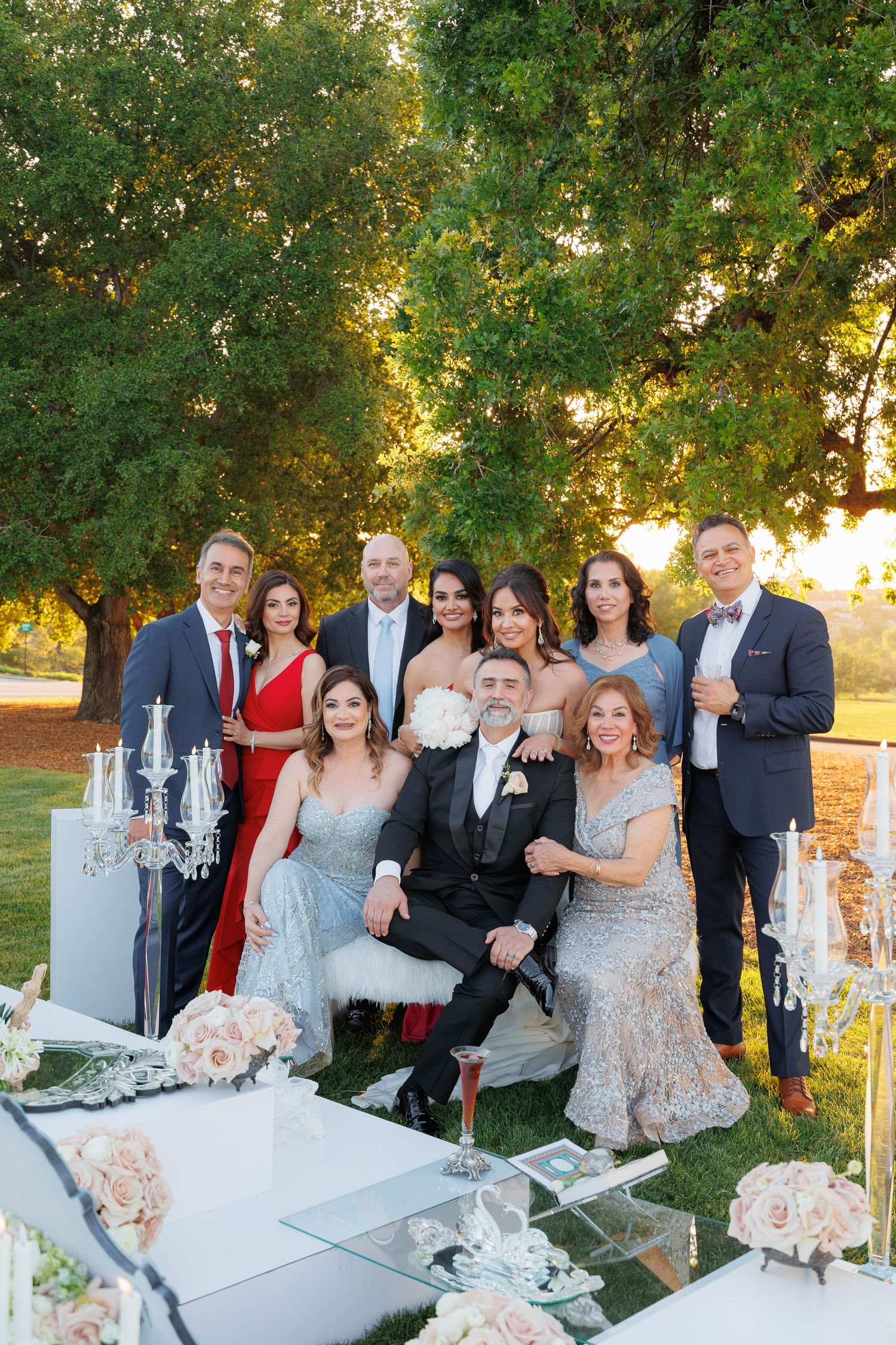 Wedding guests gather around an elegant white dessert table decorated with crystal candelabras at sunset outdoors.