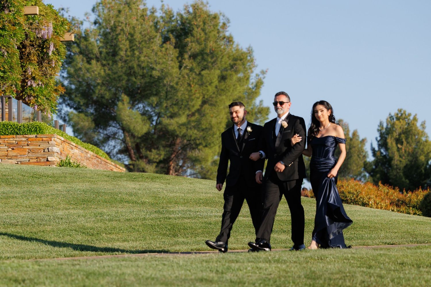 People in formal attire sitting on a grassy hill during a sunny outdoor gathering.