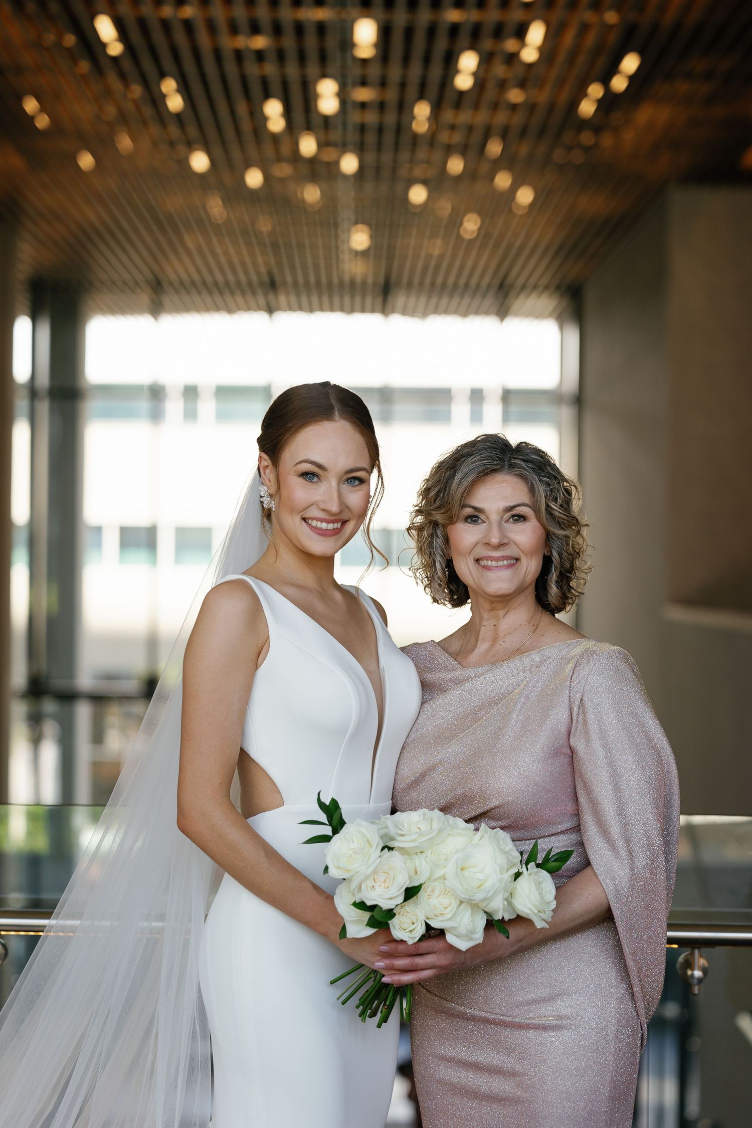 A bride in a white gown and companion in blush pink dress share joyful moments in an elegant indoor venue.