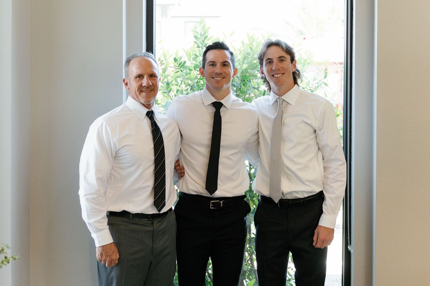 A group of people in white dress shirts and black ties stand together in a bright room near windows.