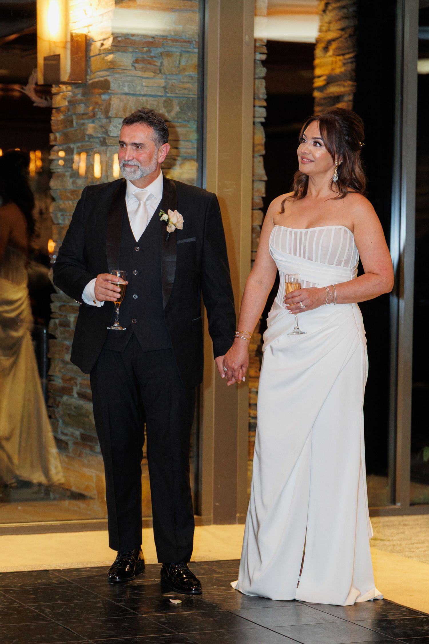 A couple in formal wedding attire holds champagne glasses while walking together during their reception celebration.