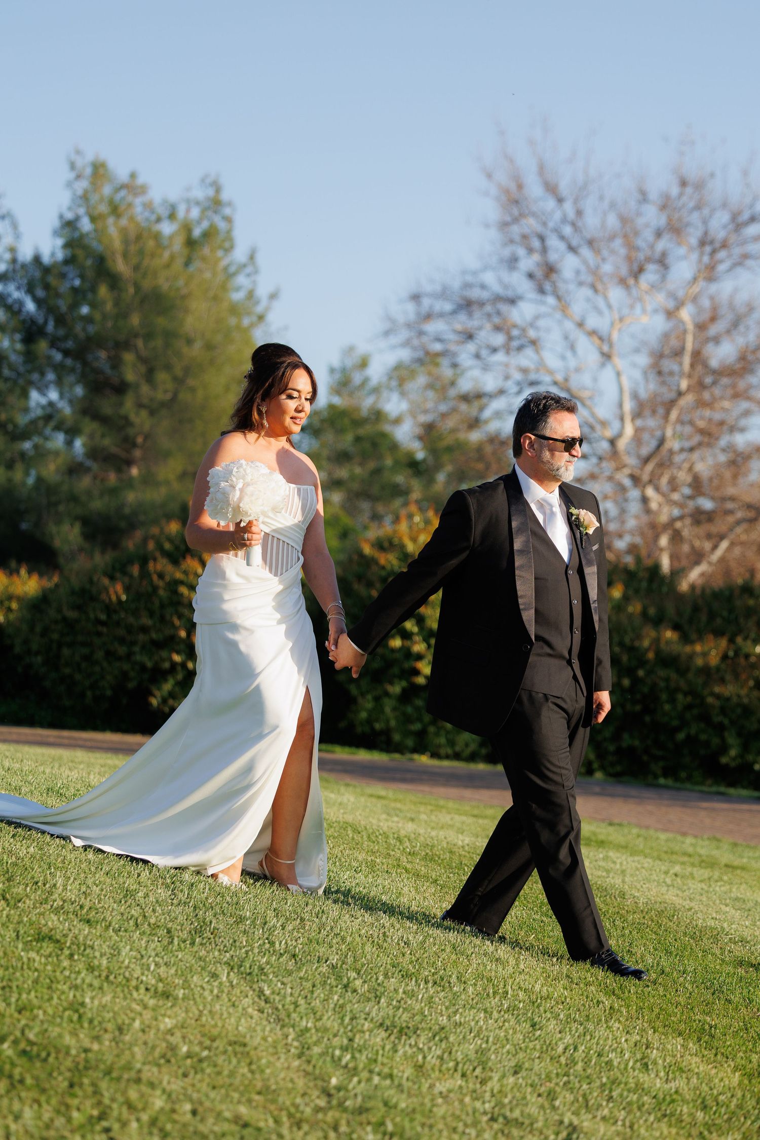 A couple walks together on a grassy lawn during their wedding, with the bride in white and groom in black formal attire.