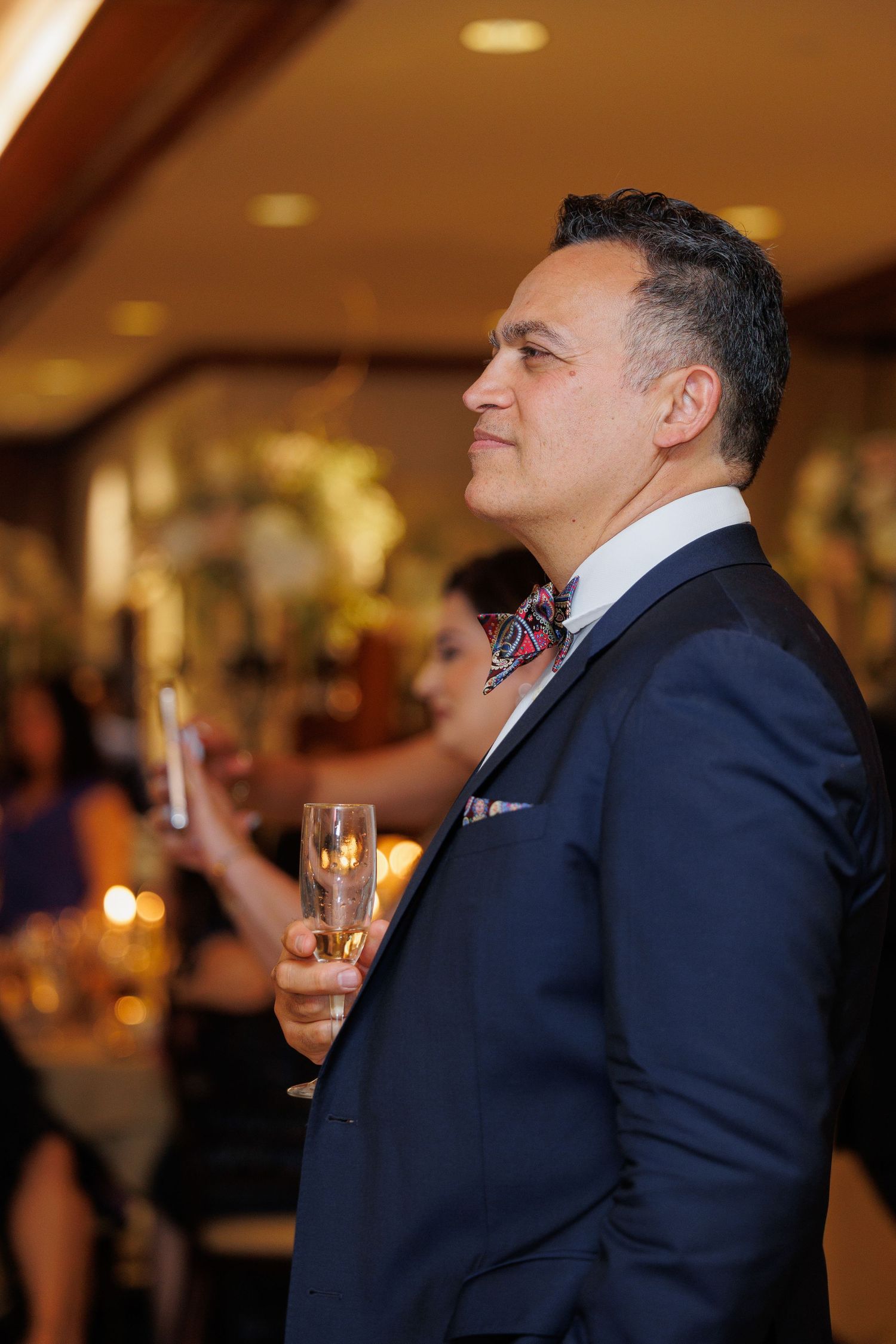 A person in a navy suit holds a champagne glass while smiling at a formal event.