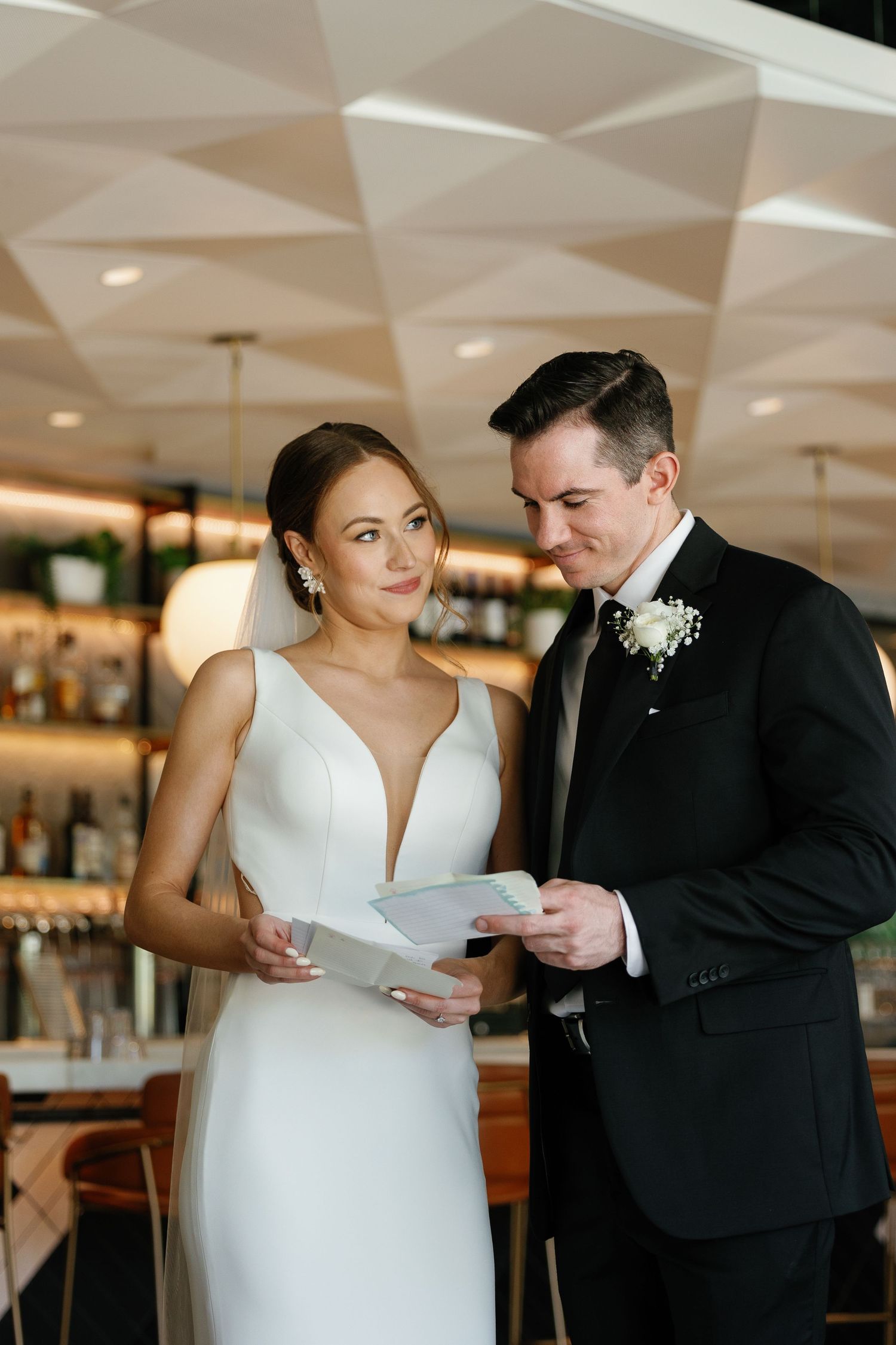 A couple in wedding attire reads a document together at an elegant bar with geometric ceiling details.