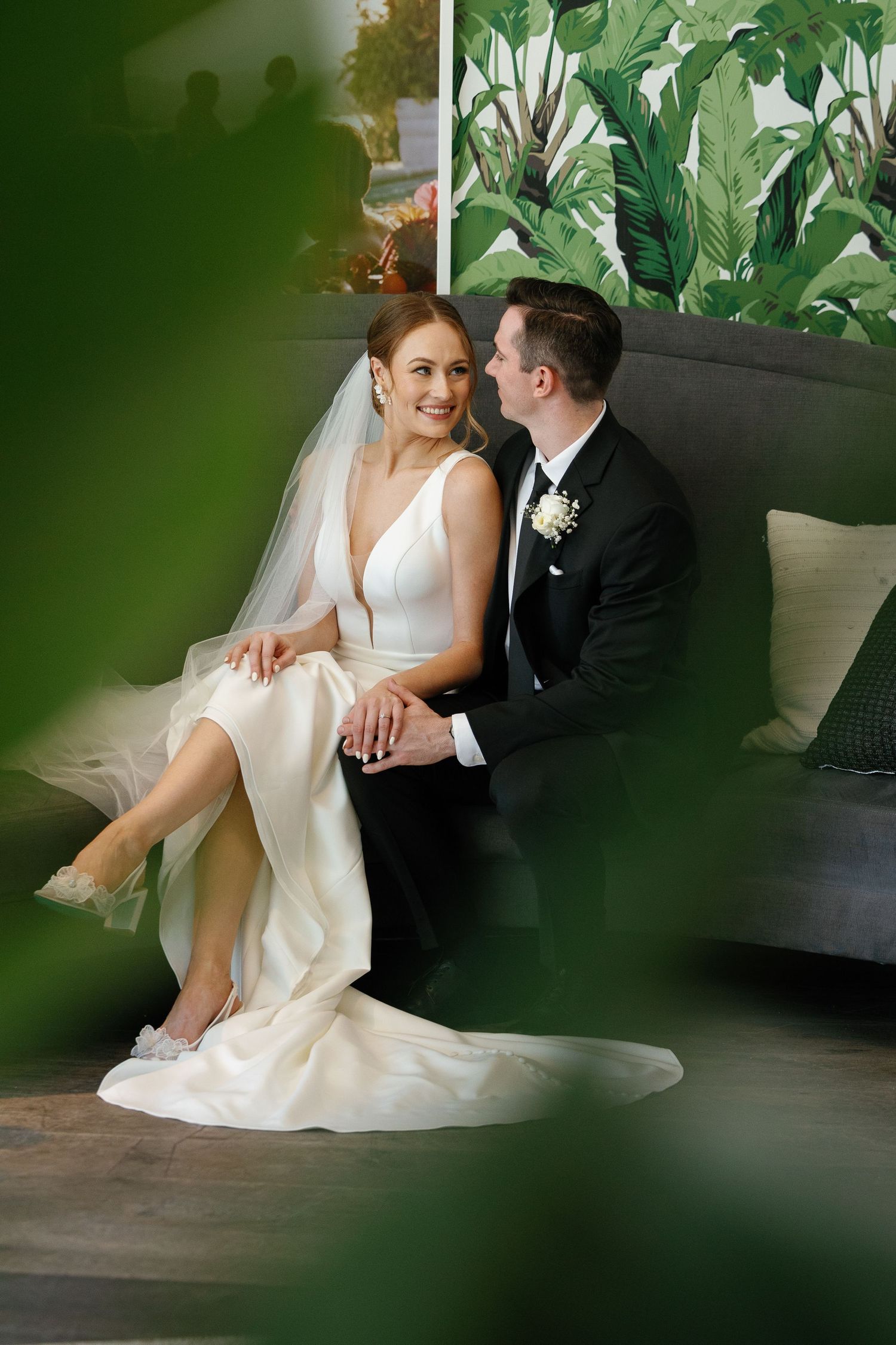 Romantic wedding couple sitting together on dark floor with green foliage framing the intimate scene through window.