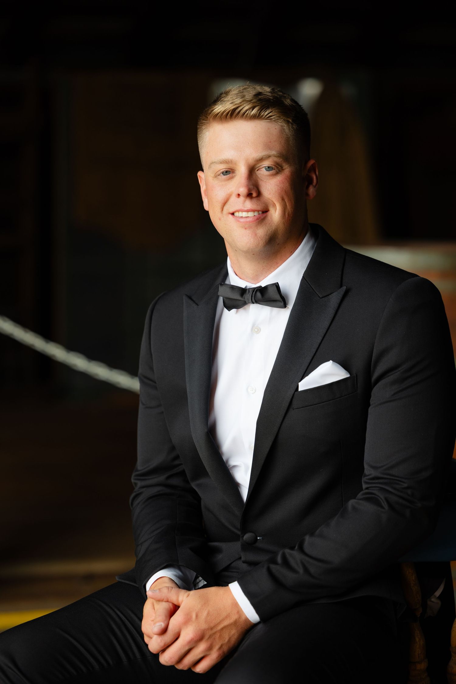 A person in a classic black tuxedo and white bow tie poses for formal portraits against a dark background.