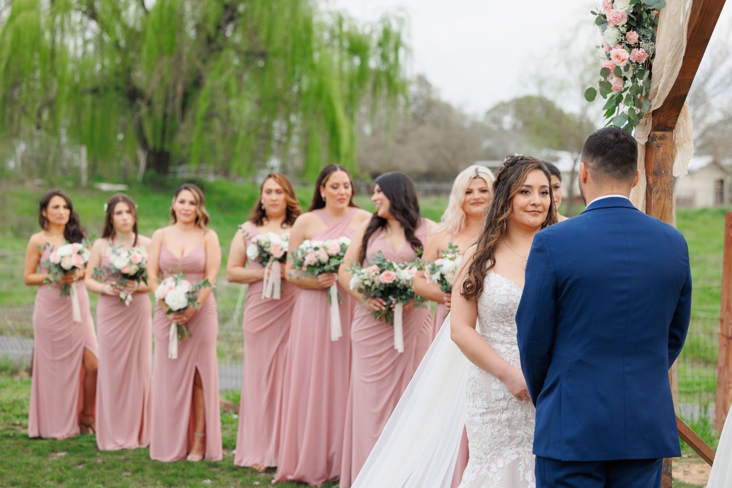 A wedding ceremony outdoors with bridesmaids in pink dresses standing in a row under willow trees.