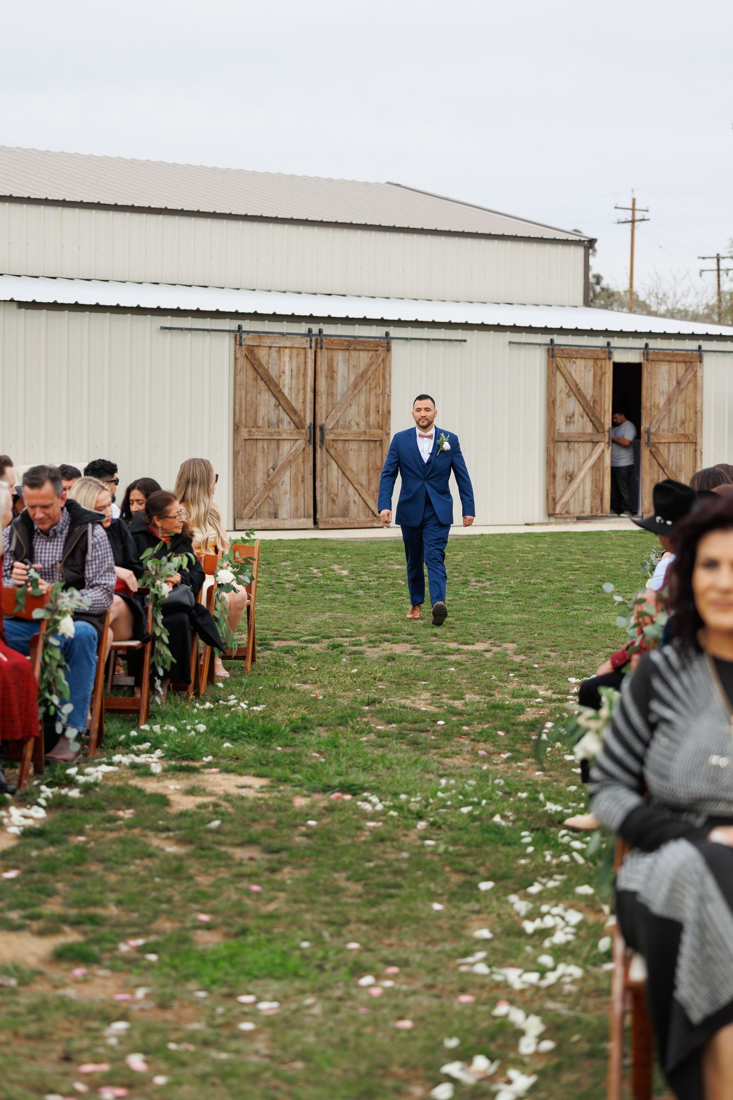 People gather on a grassy field near a large barn building with wooden doors on an overcast day.