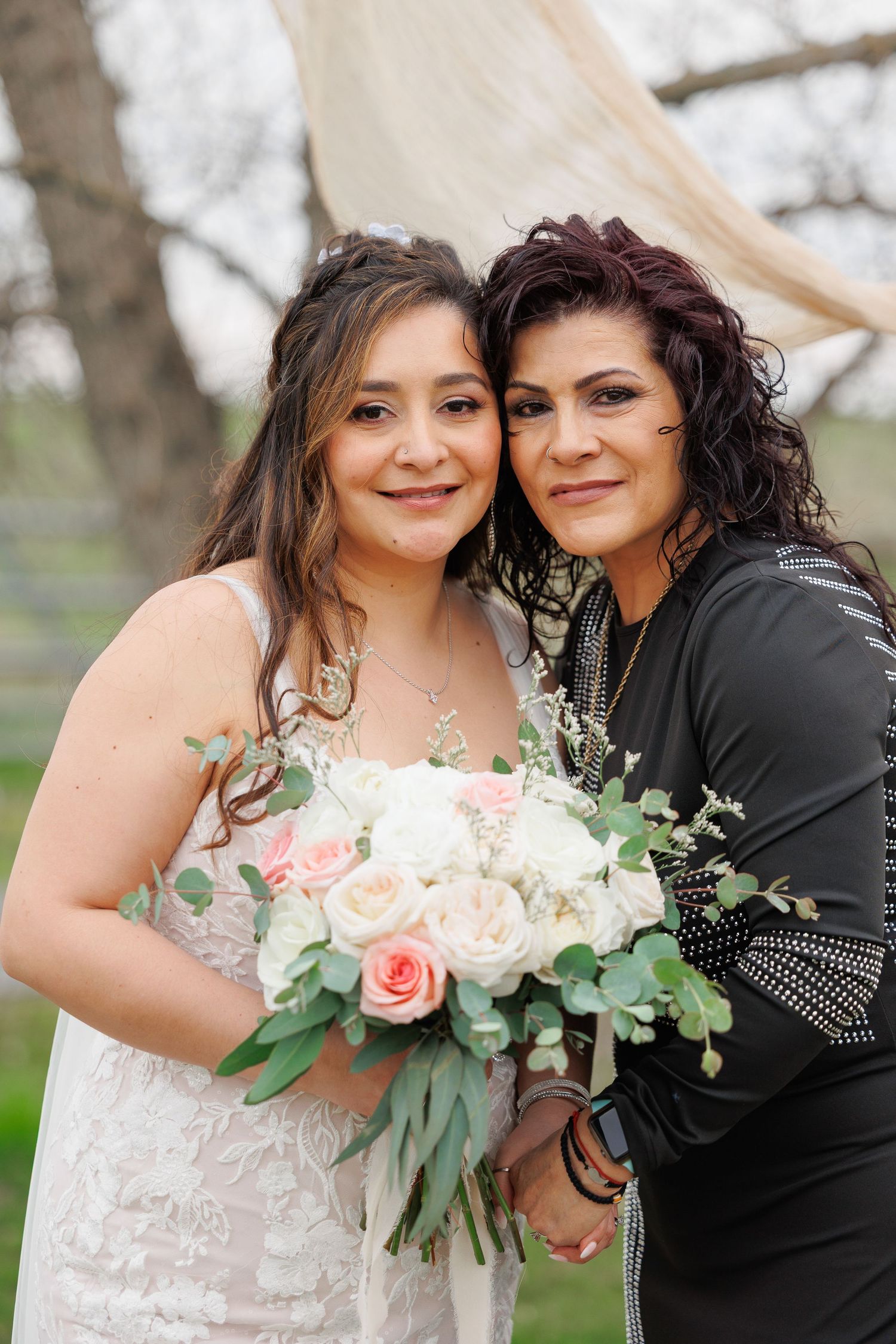 Two people pose together at an outdoor wedding, with one in a white dress holding a pink and white floral bouquet.
