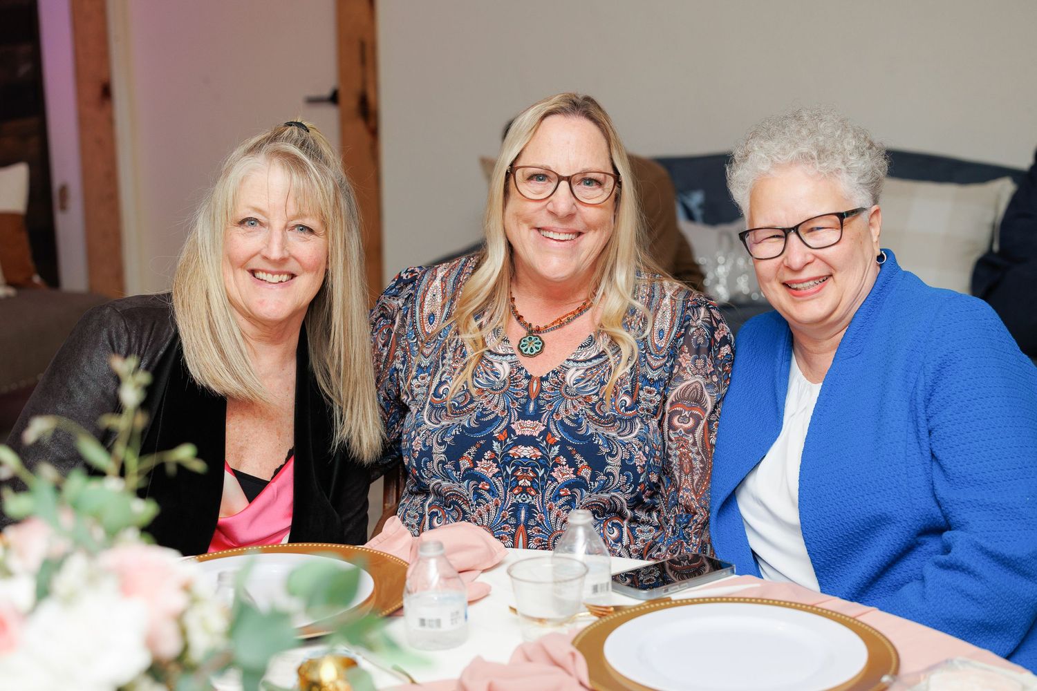 Three people smile warmly while sitting together at a dining table with place settings and floral decor.