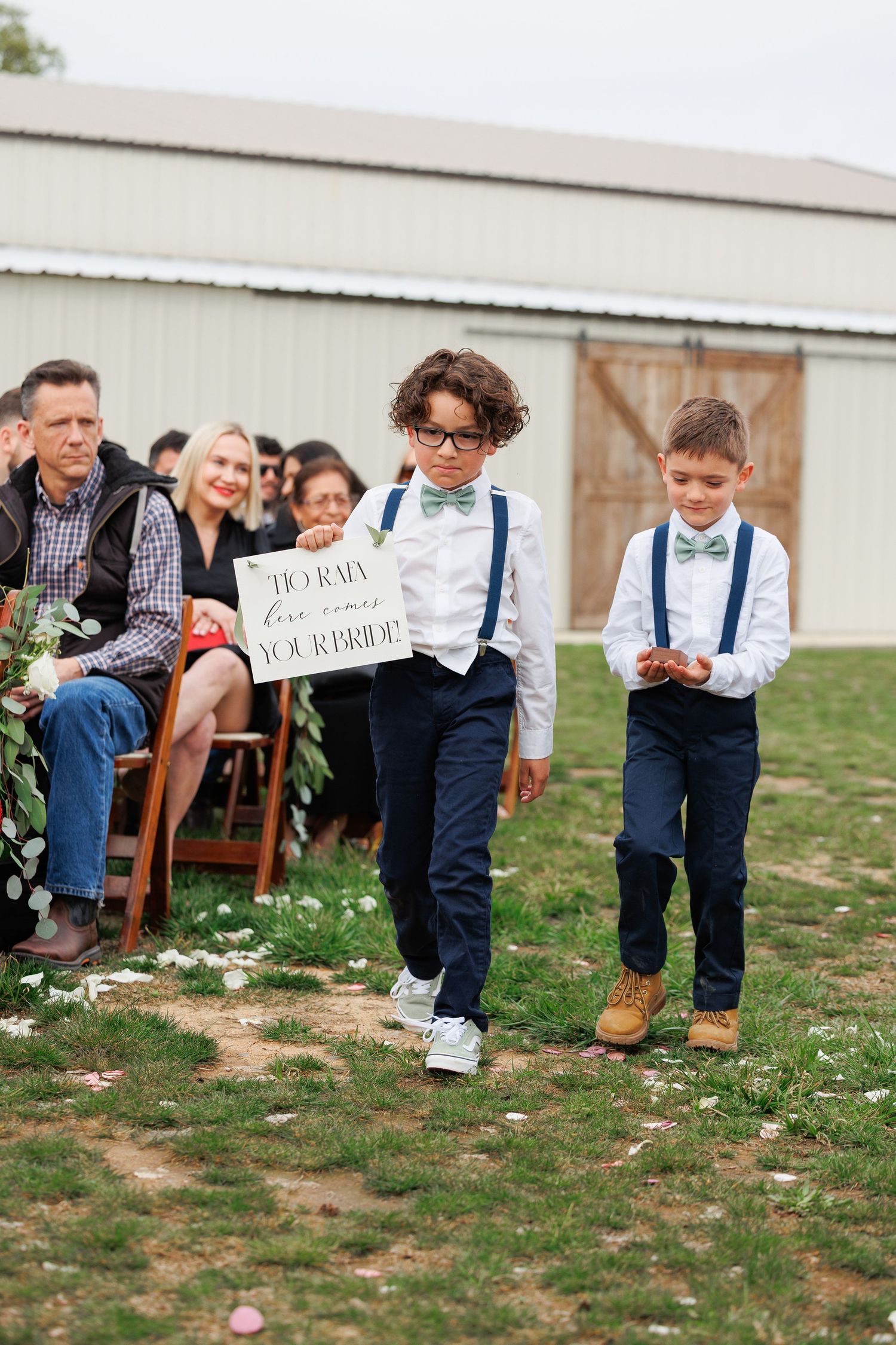 Children in suspenders and dress clothes play on the grass during an outdoor wedding celebration.