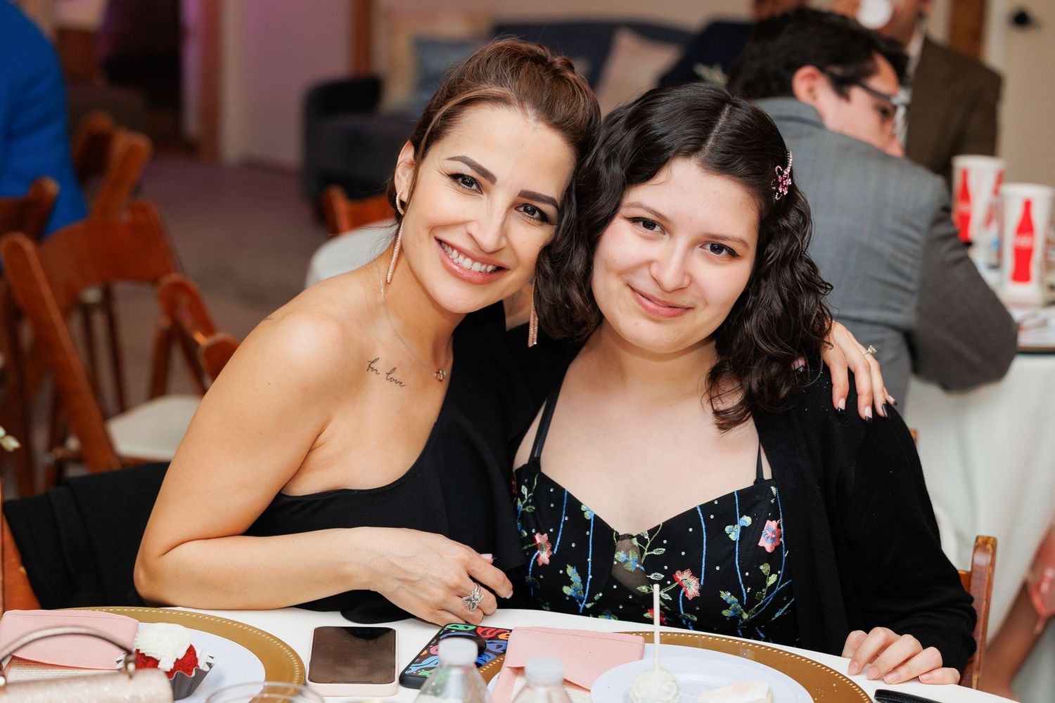 Two people in black attire share a warm smile while sitting together at a dining table during an event.
