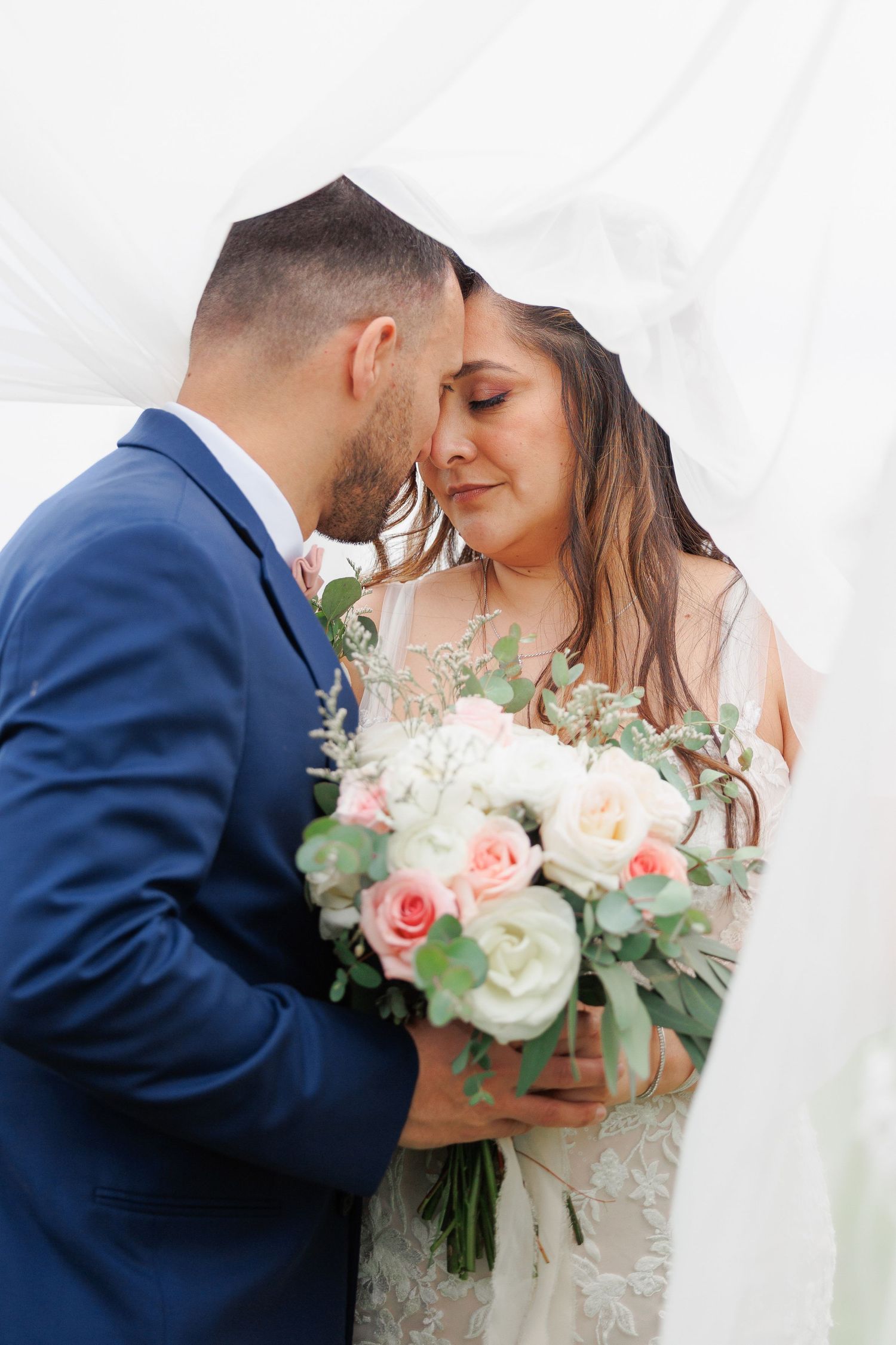A romantic wedding moment with a bride in white lace holding a pink and white rose bouquet next to a groom in navy blue suit.