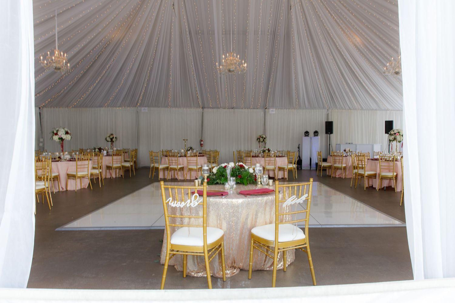 Interior view of an elegant white wedding tent with round tables and gold chairs set up for a formal reception event.