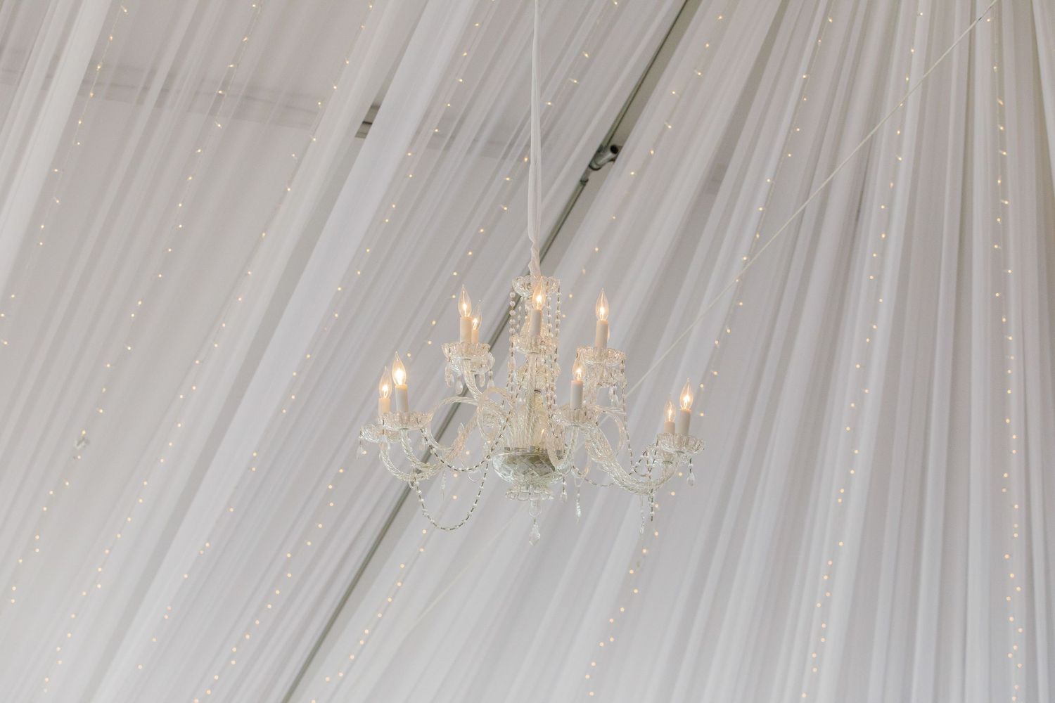 Elegant white fabric drapes and crystal chandeliers create a luxurious wedding reception ceiling design.