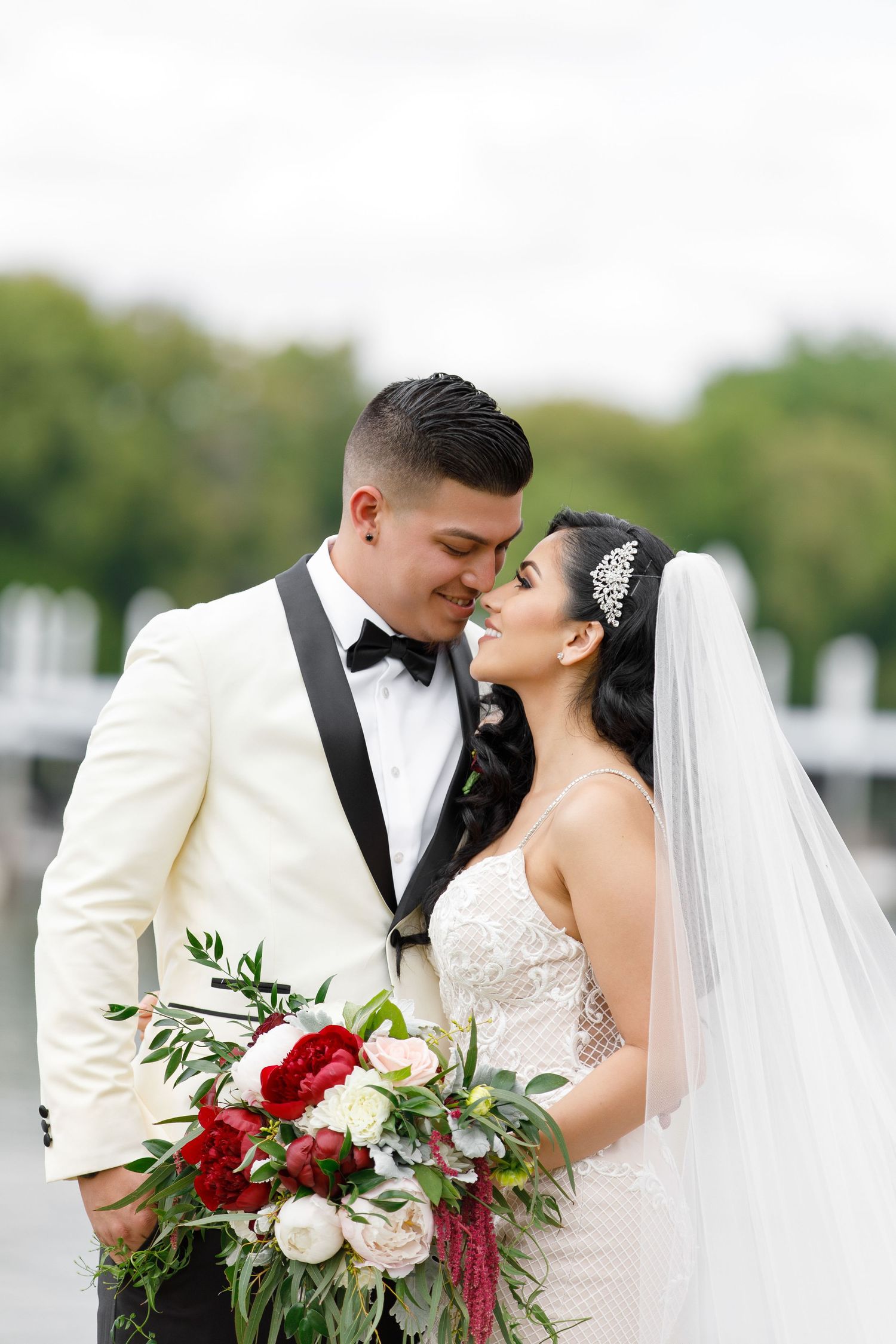 Elegant wedding couple in white tuxedo and lace dress share intimate moment with stunning red and white bouquet.