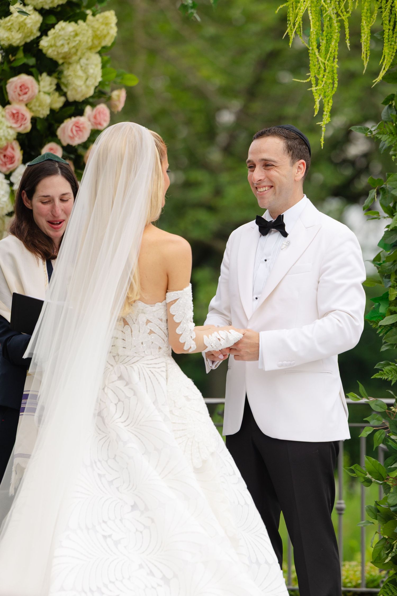 A bride and groom exchange rings during an outdoor garden wedding ceremony surrounded by floral arrangements.