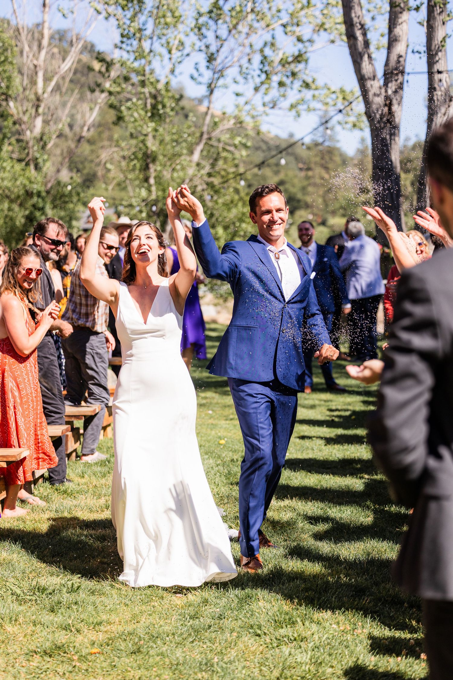 Wedding guests celebrate and cheer outdoors on a sunny day as newlyweds walk through a grassy area lined with trees.